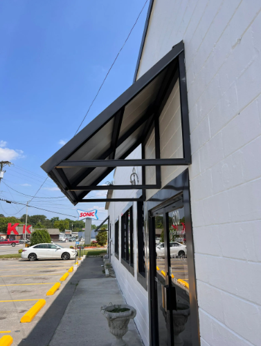 Black awning over the door of a white building with black trim, sidewalk, and cars parked on a sunny day.