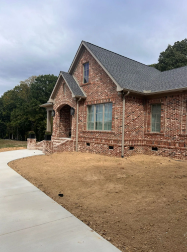Brick house with a gray roof, green-framed windows, and a concrete walkway on a cloudy day.