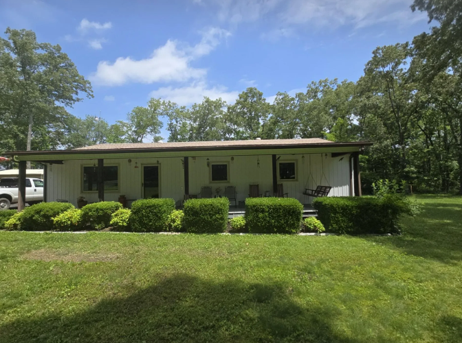 White bungalow house with green lawn and bushes, trees in background, blue sky.