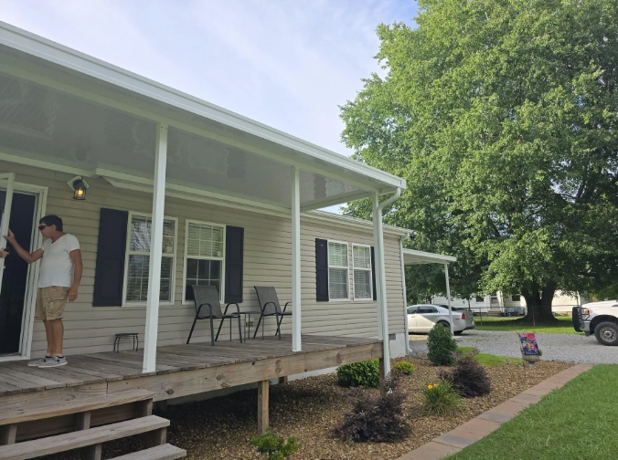 Man on porch of a beige house, opening door. Black shutters, chairs, large tree, sunny.