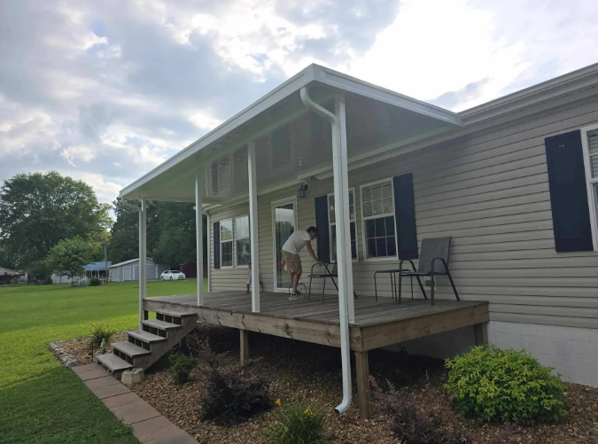 Person on porch of a house with a covered roof, setting up furniture. Wooden steps, green lawn.