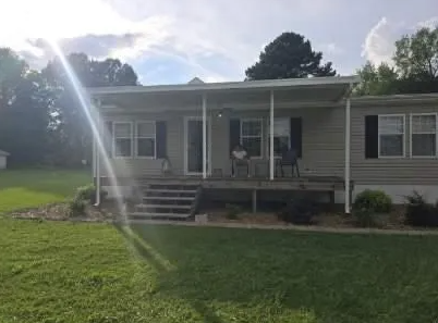 Tan house with porch, steps, and awning. Green lawn in the foreground. Cloudy sky.