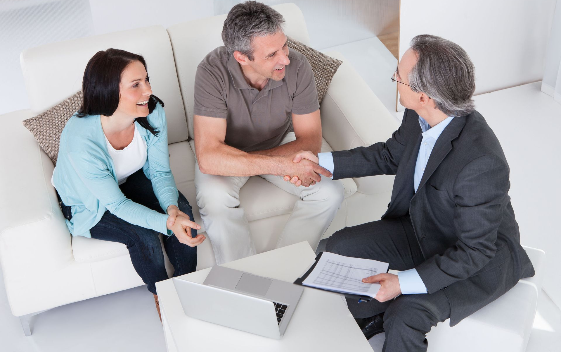 Three people in a meeting, shaking hands and discussing papers in a bright office lounge.
