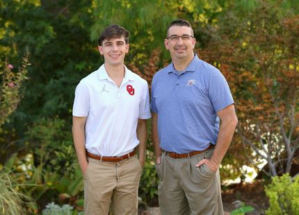 Two men standing outdoors, smiling in front of greenery, one in a white polo and one in a blue striped shirt