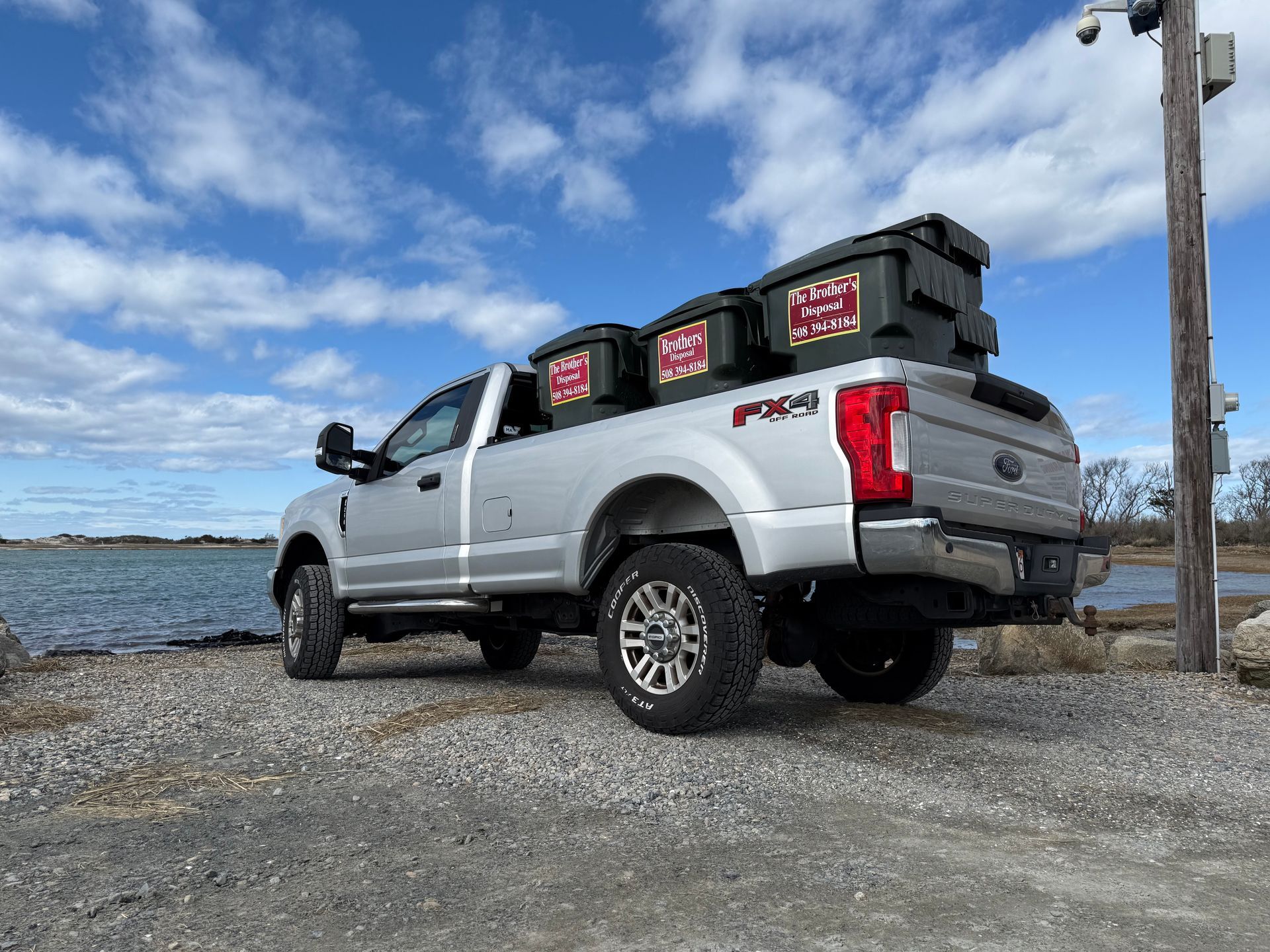Red pickup truck carrying three green trash bins; setting is a residential area.