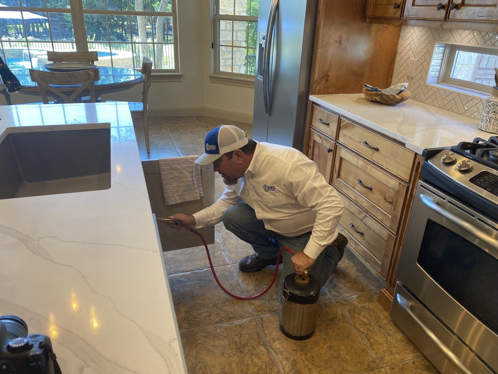 Pest control worker sprays a kitchen countertop. He is wearing a uniform and a baseball cap, kneeling with a sprayer.