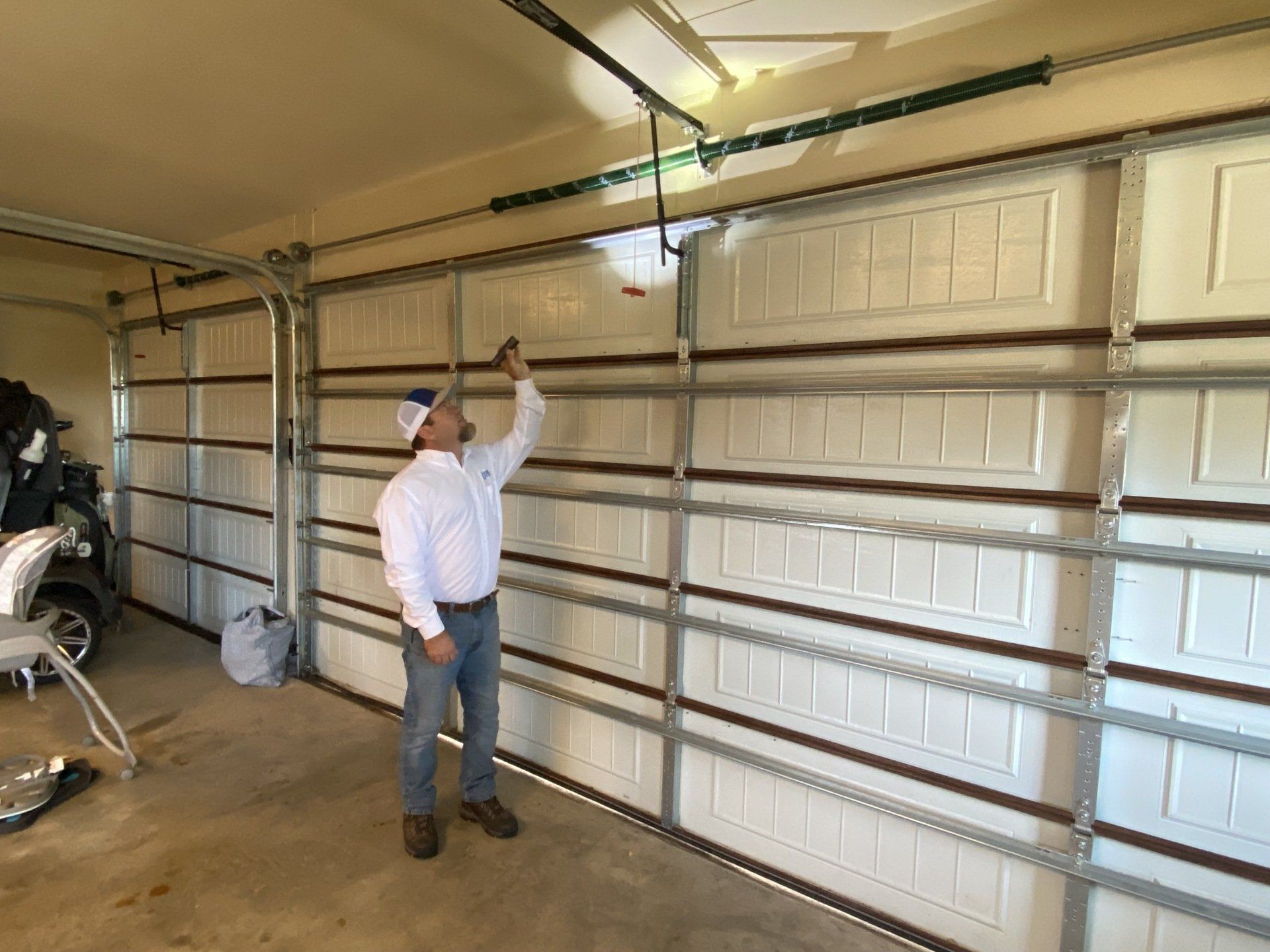 A man inspects a white garage door. He wears a button-down shirt, jeans, and a hat in a garage with concrete floors.