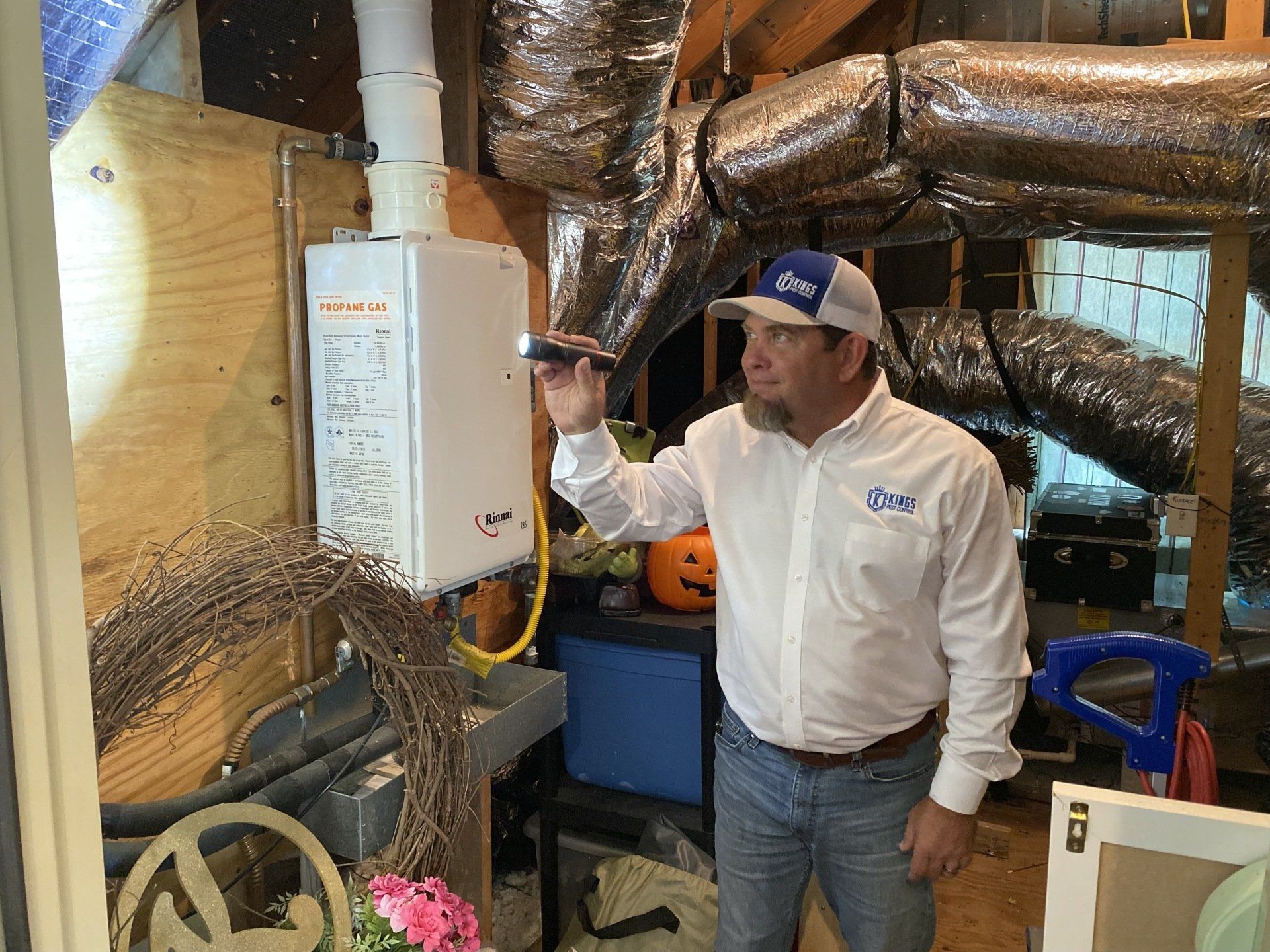 A man in a white shirt and baseball cap examines a water heater with a flashlight in an attic.