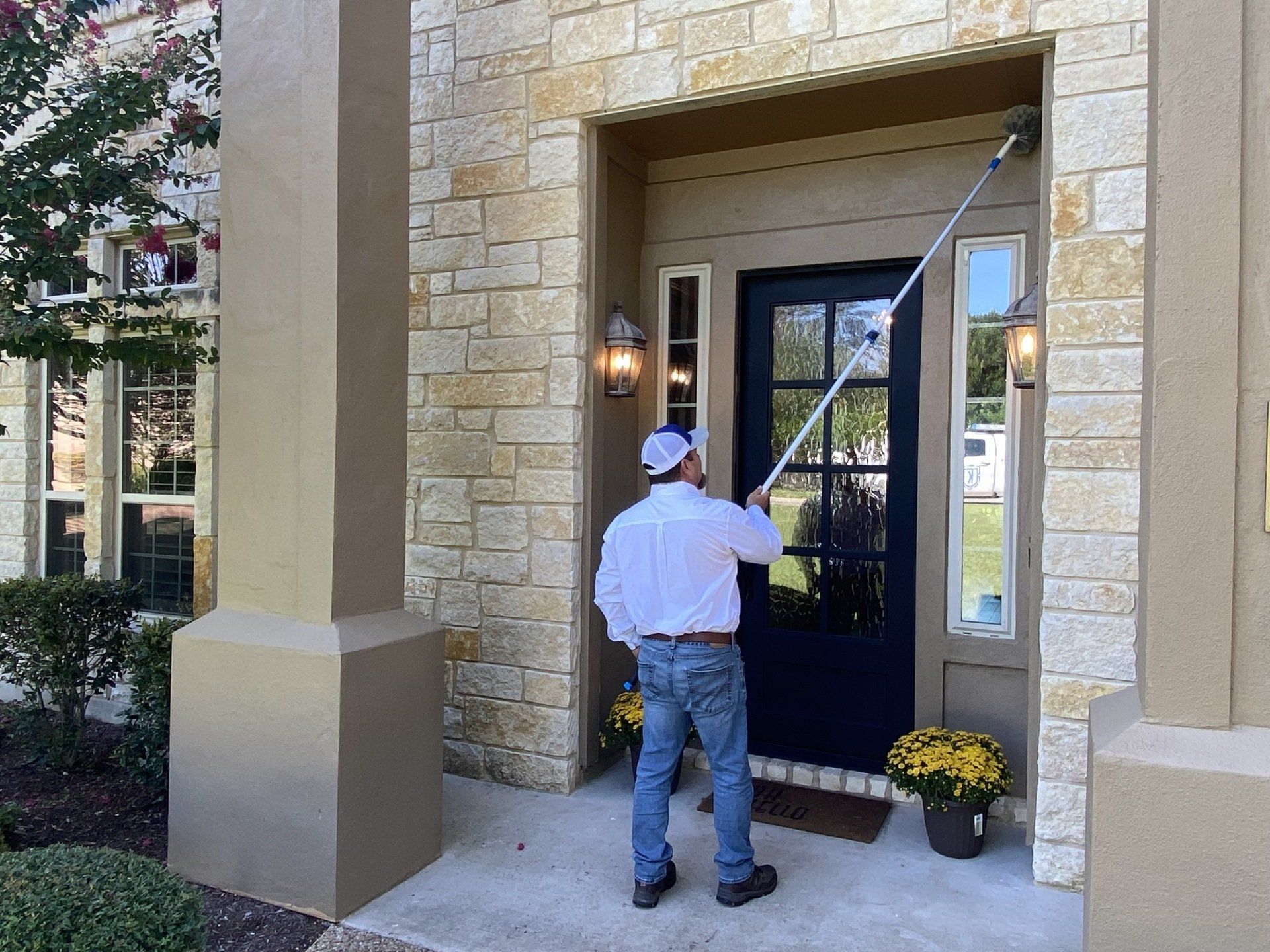 A man cleaning a front door with a long-handled tool. He is wearing a white cap and shirt, standing in front of a stone-clad entrance.
