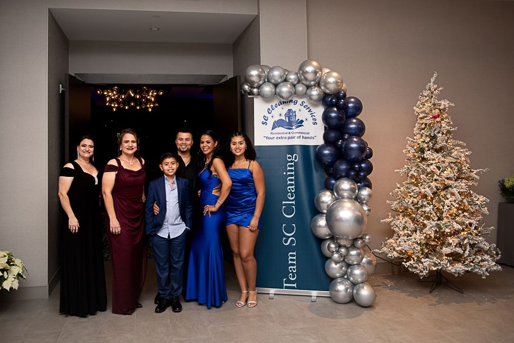 Group poses at a festive event: formal attire, balloon arch, Christmas tree.