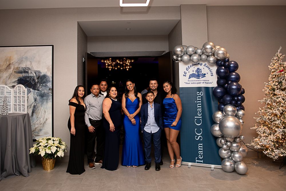 Group of ten people in formal wear pose for a photo at an event. Silver and blue balloons arch over a banner.