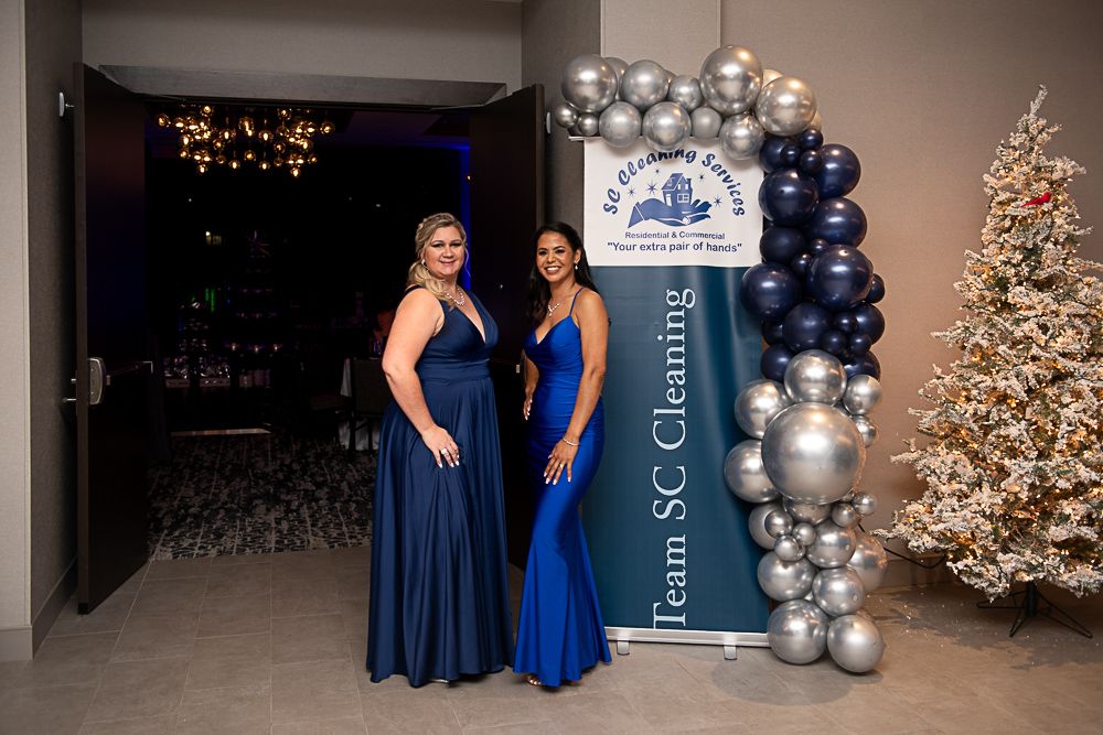 Two women in formal gowns pose by a banner at an event with balloon arch and Christmas tree.