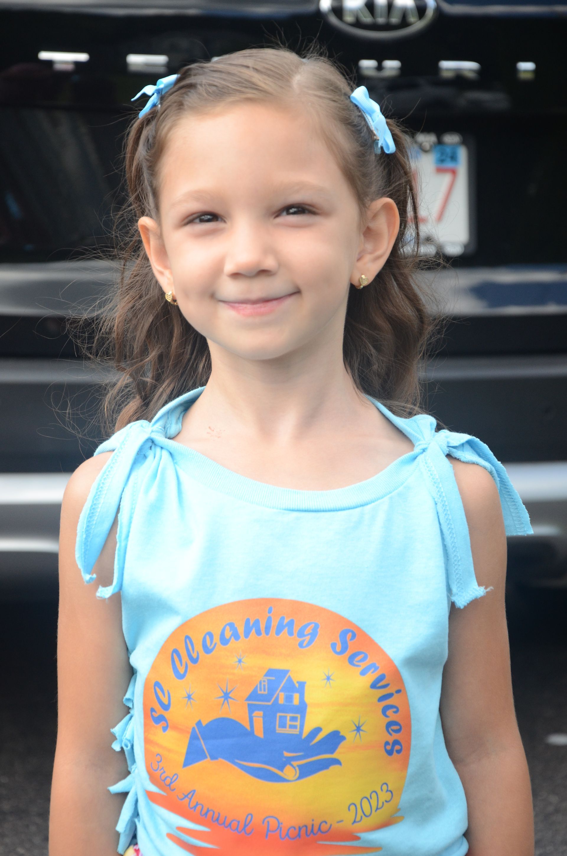 Girl with brown hair and blue bows smiles, wearing a light blue shirt with an orange and yellow logo.