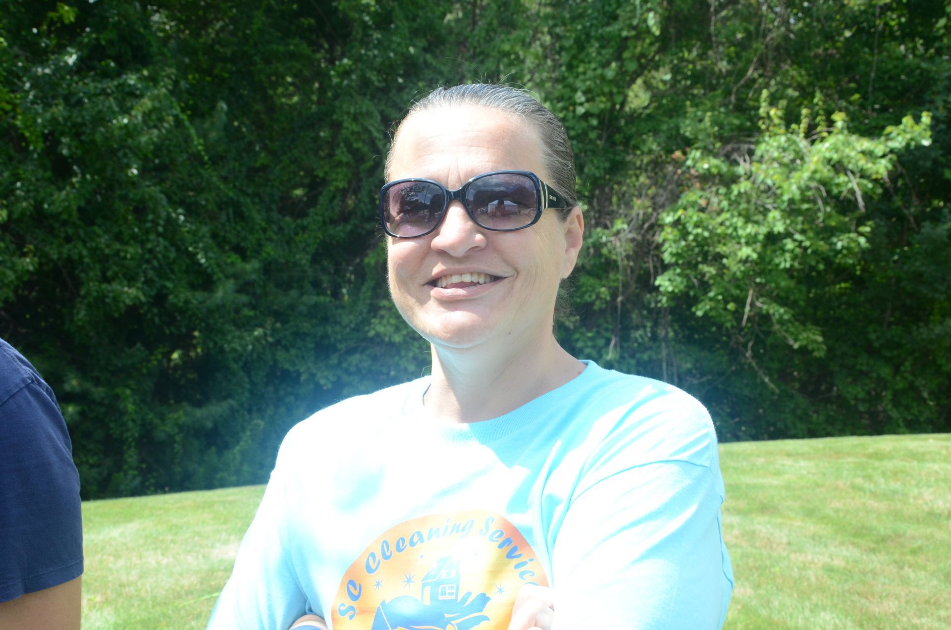 Woman with sunglasses smiles outside, wearing a light blue shirt. Green trees in the background.