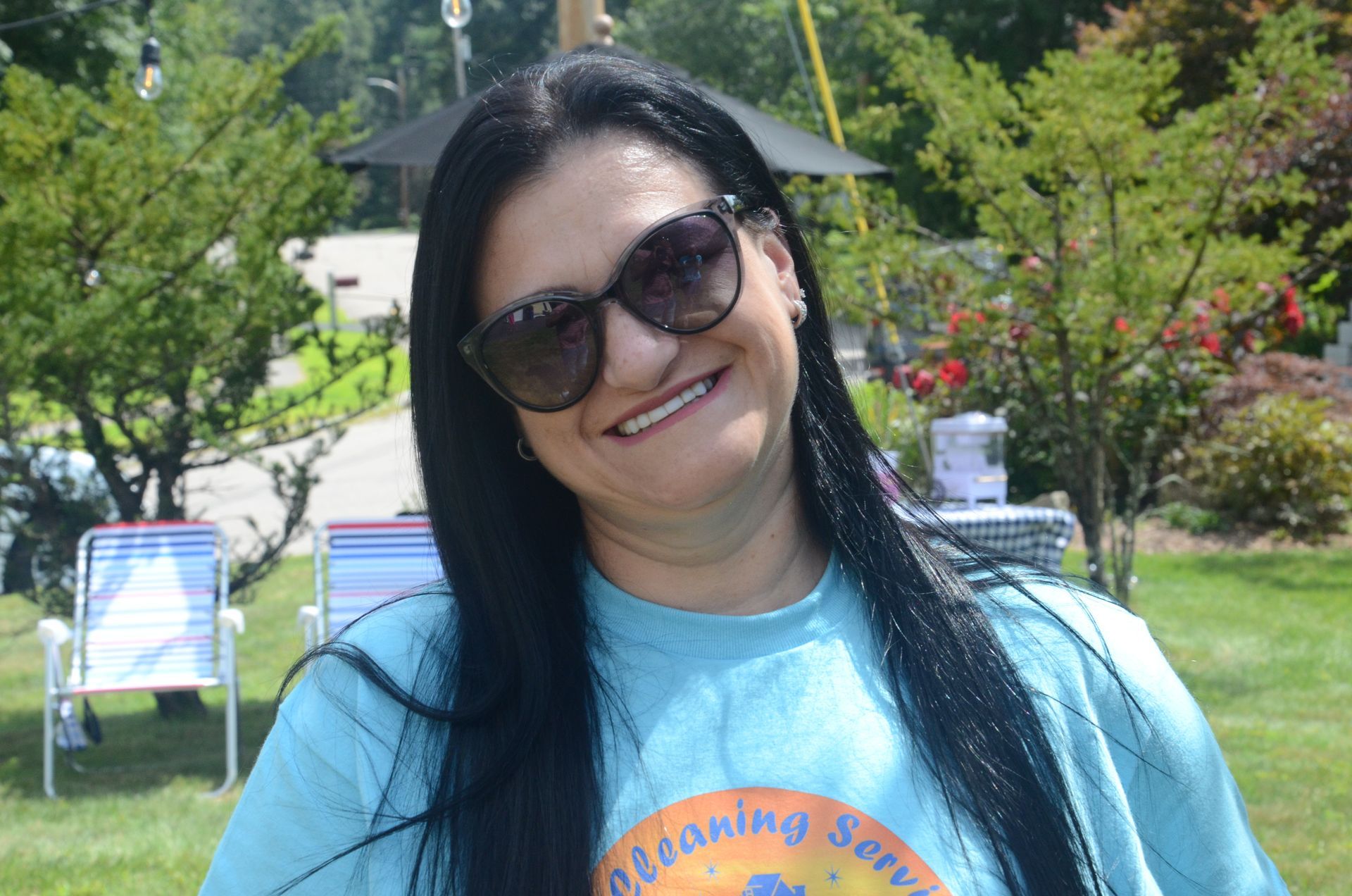 Woman with sunglasses smiles outdoors, wearing a teal shirt. Green grass and trees are visible in the background.