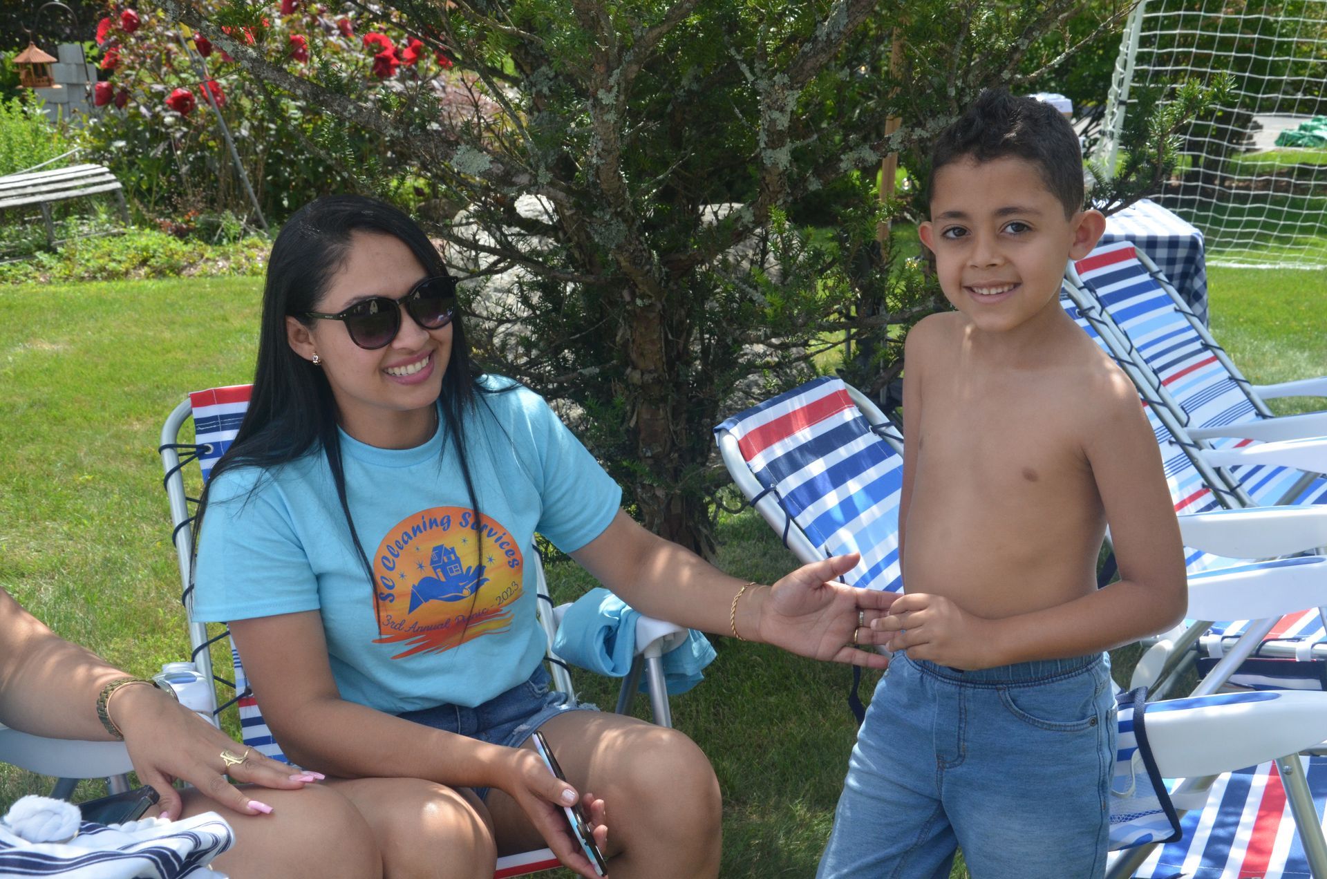 Woman in sunglasses smiles at a boy. They are outdoors, sitting in lawn chairs, with green grass in the background.