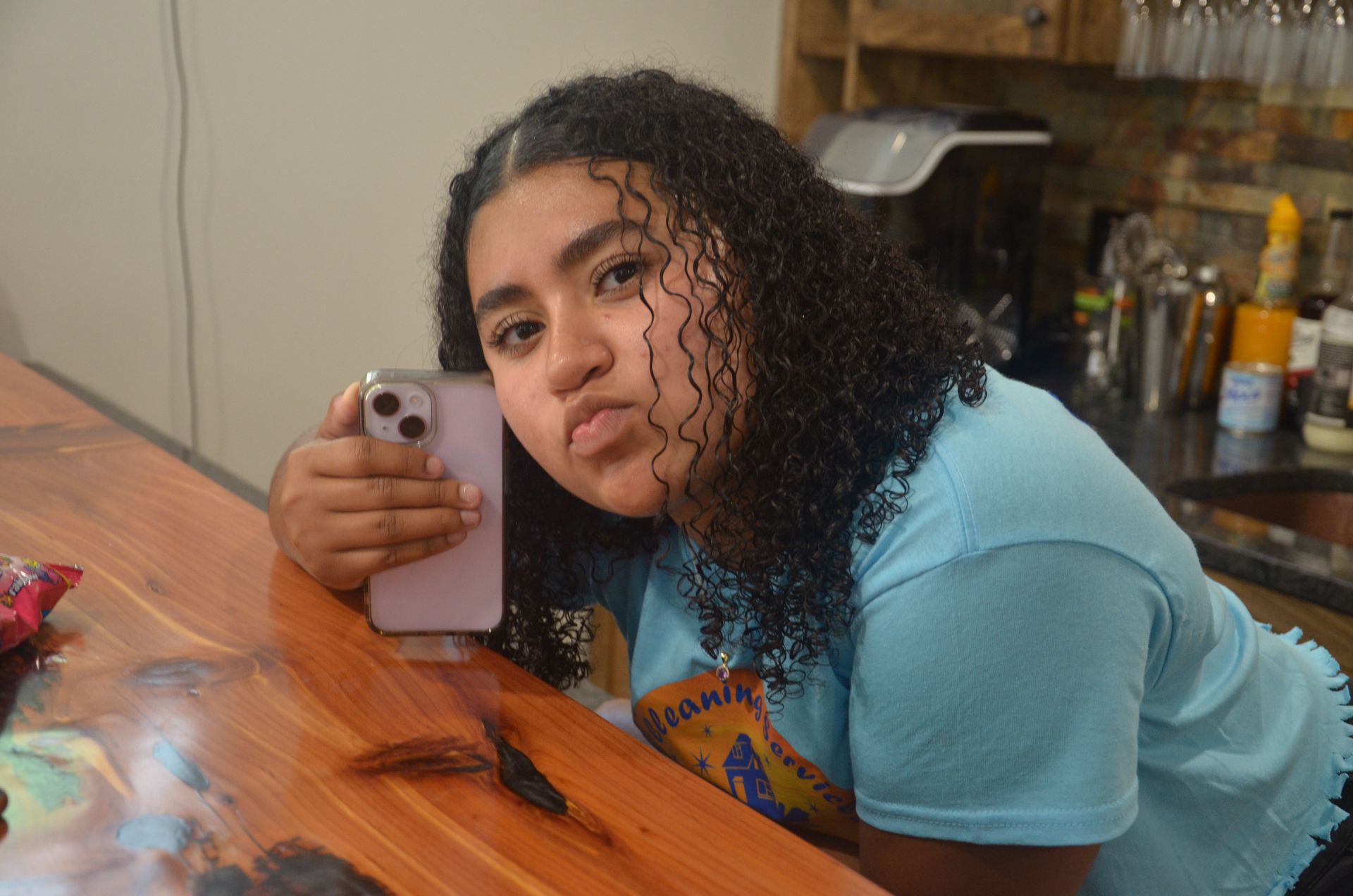 A young woman with curly hair takes a selfie, leaning on a wooden bar, making a slight face.