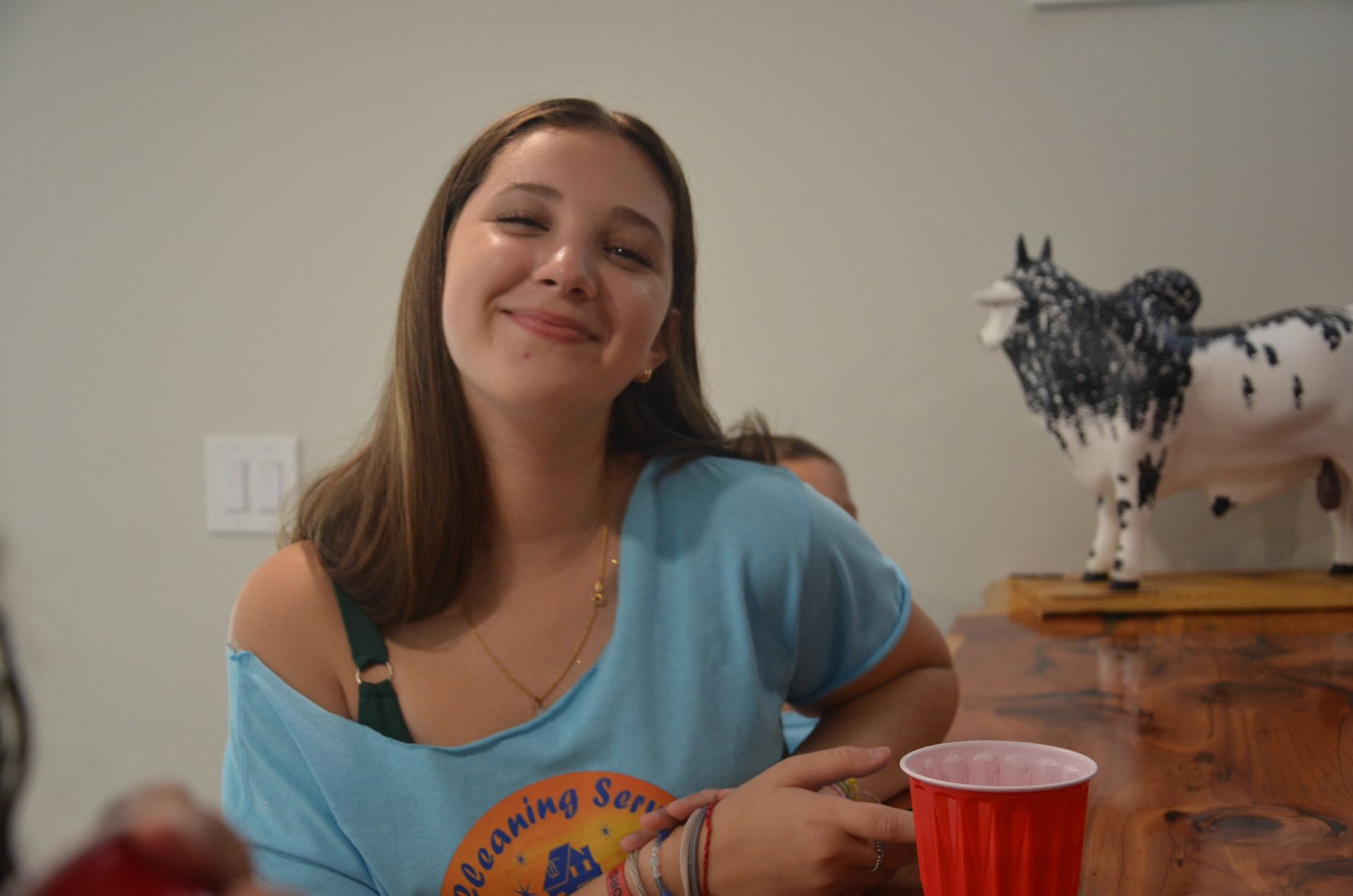 Woman smiling, wearing blue shirt, holding red cup, next to cow statue, indoors.