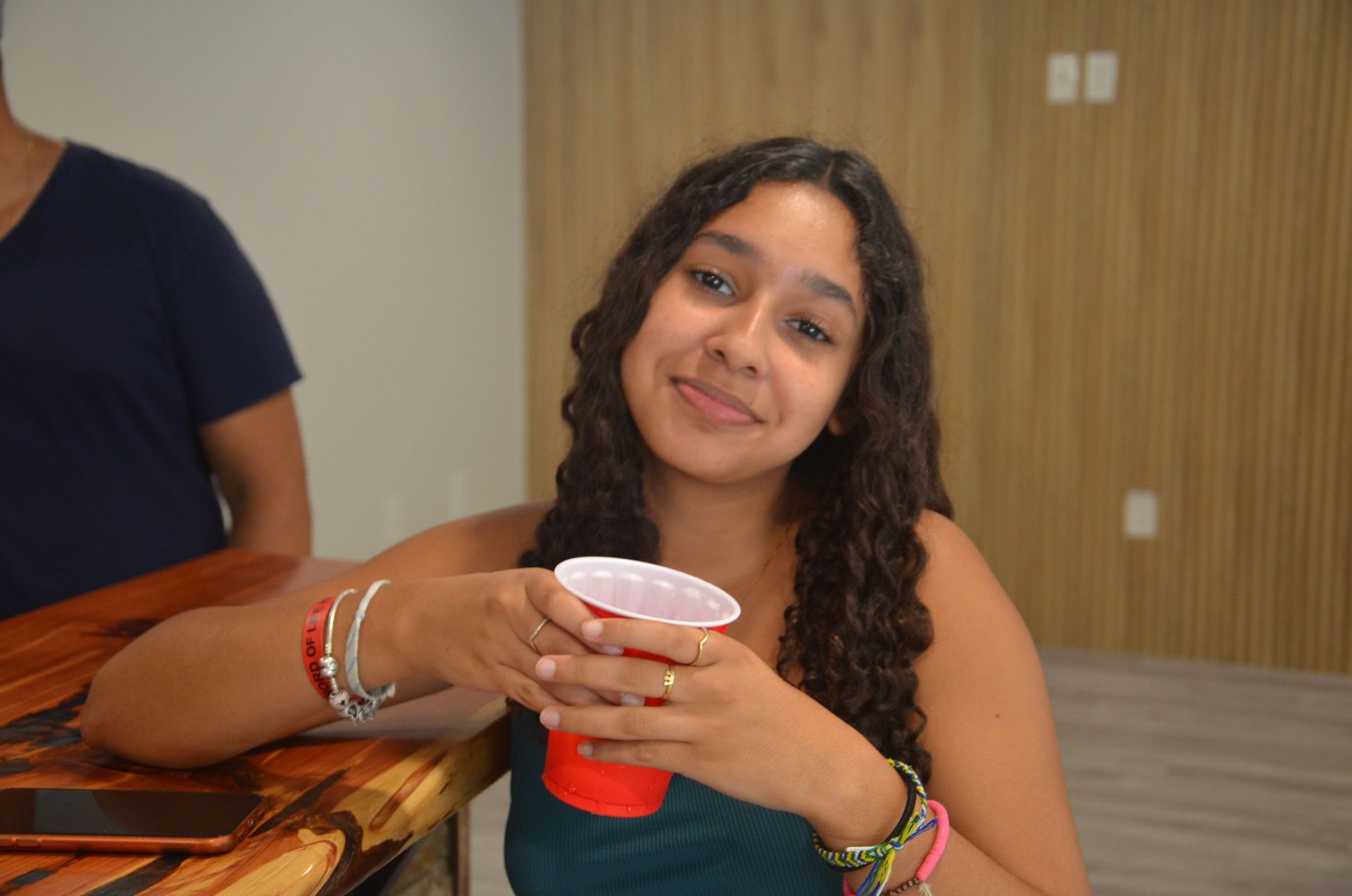 Woman with curly hair smiles, holding a red cup at a bar.