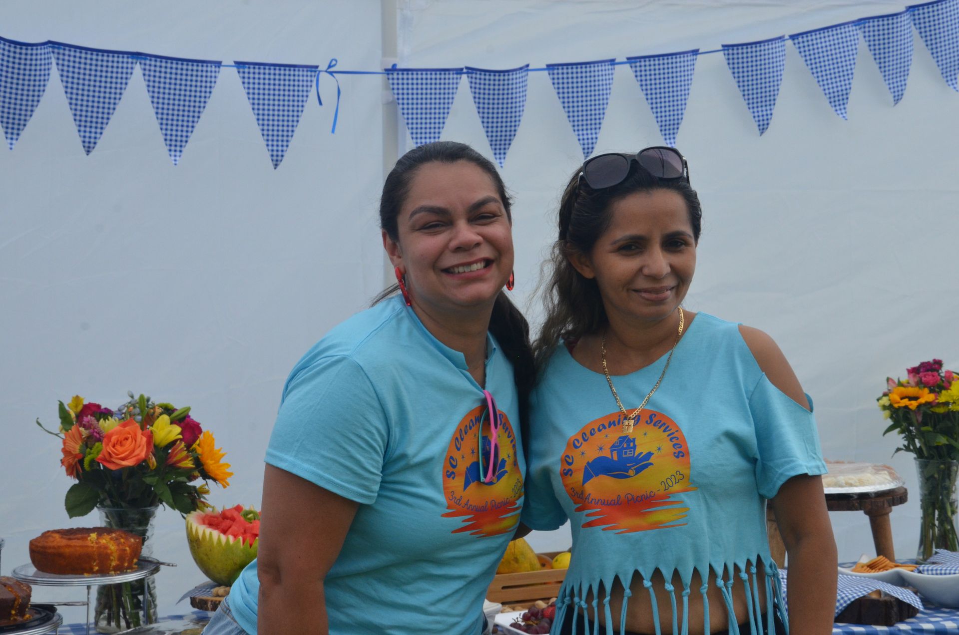 Two women in blue shirts smiling, at an outdoor event with flowers and food.