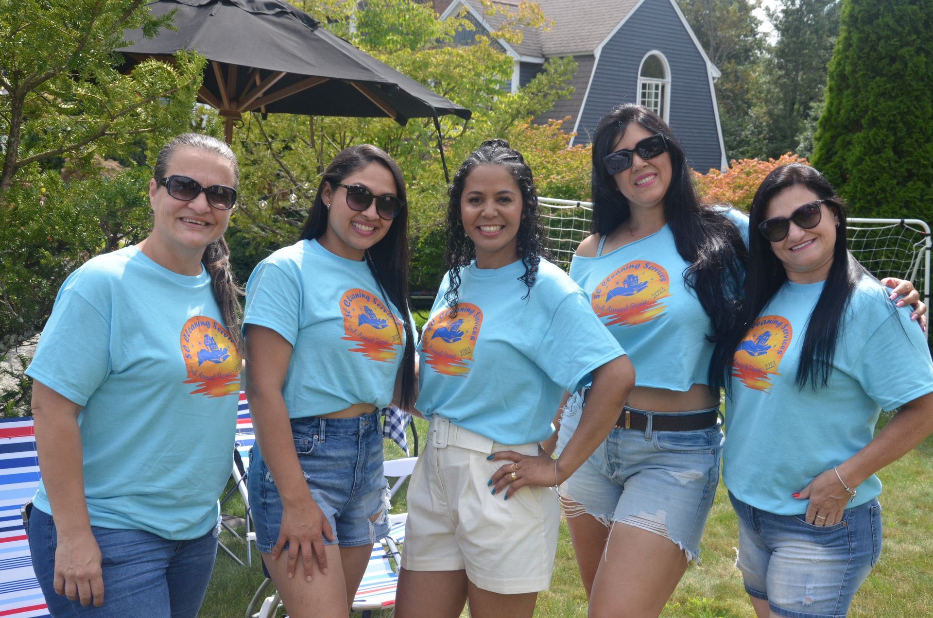 Five women in matching blue shirts and sunglasses pose in a backyard.