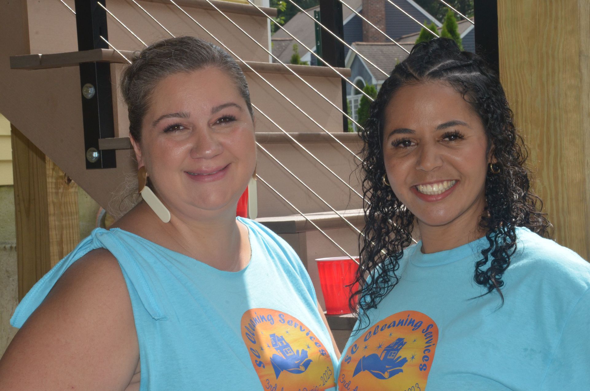 Two women smiling, wearing matching blue t-shirts. Outside, near steps.