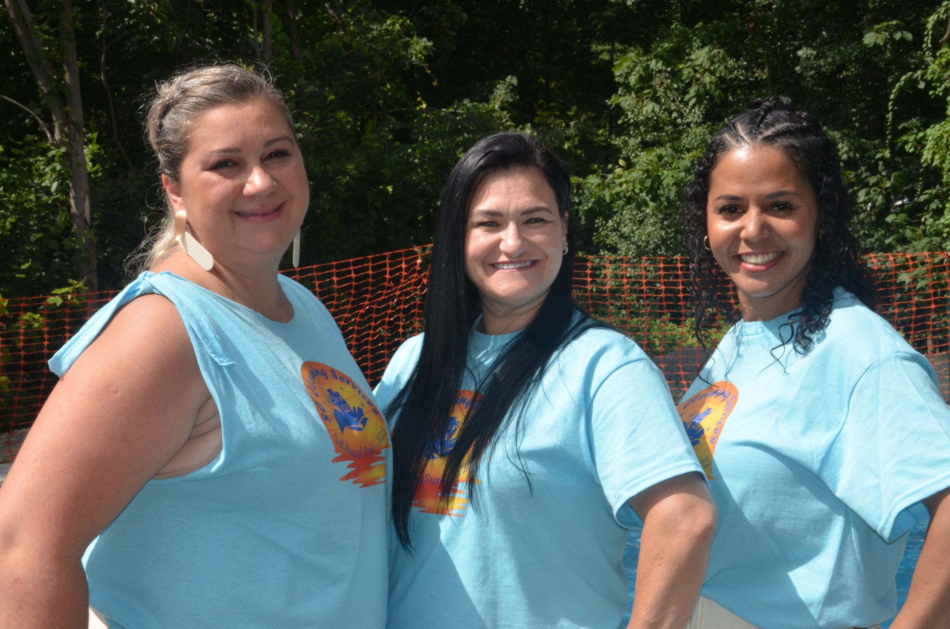 Three women in light blue t-shirts smile outdoors.