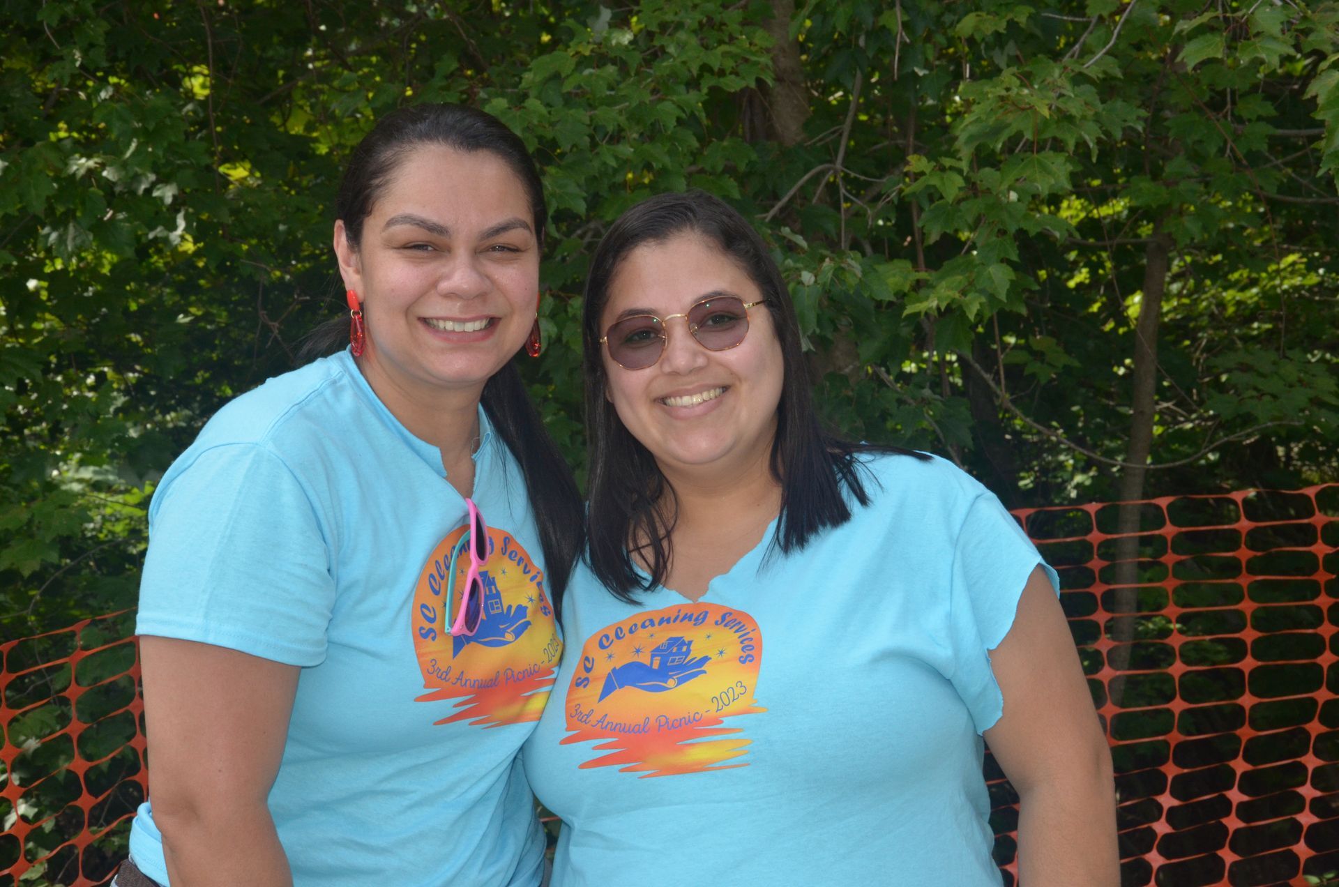 Two smiling women in blue shirts pose outdoors, near a tree and orange fencing.