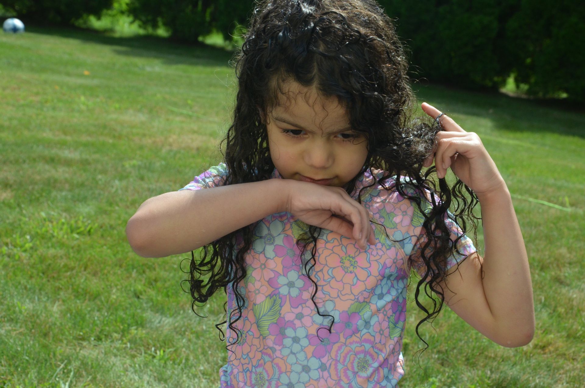 Girl with wet, curly hair in floral top touches face in grassy yard.