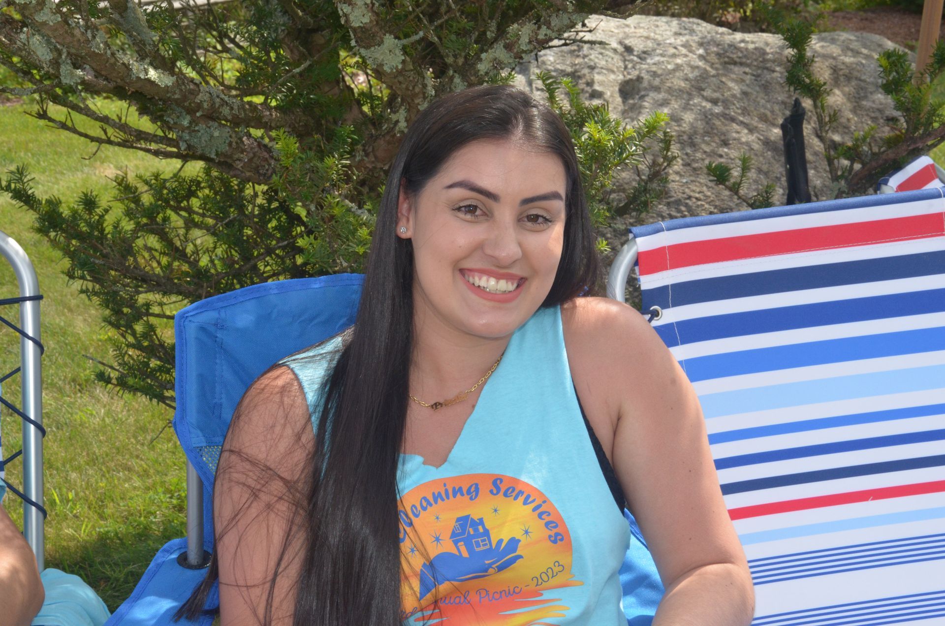 Woman with long dark hair smiles while sitting in a blue chair, wearing a turquoise shirt outdoors.