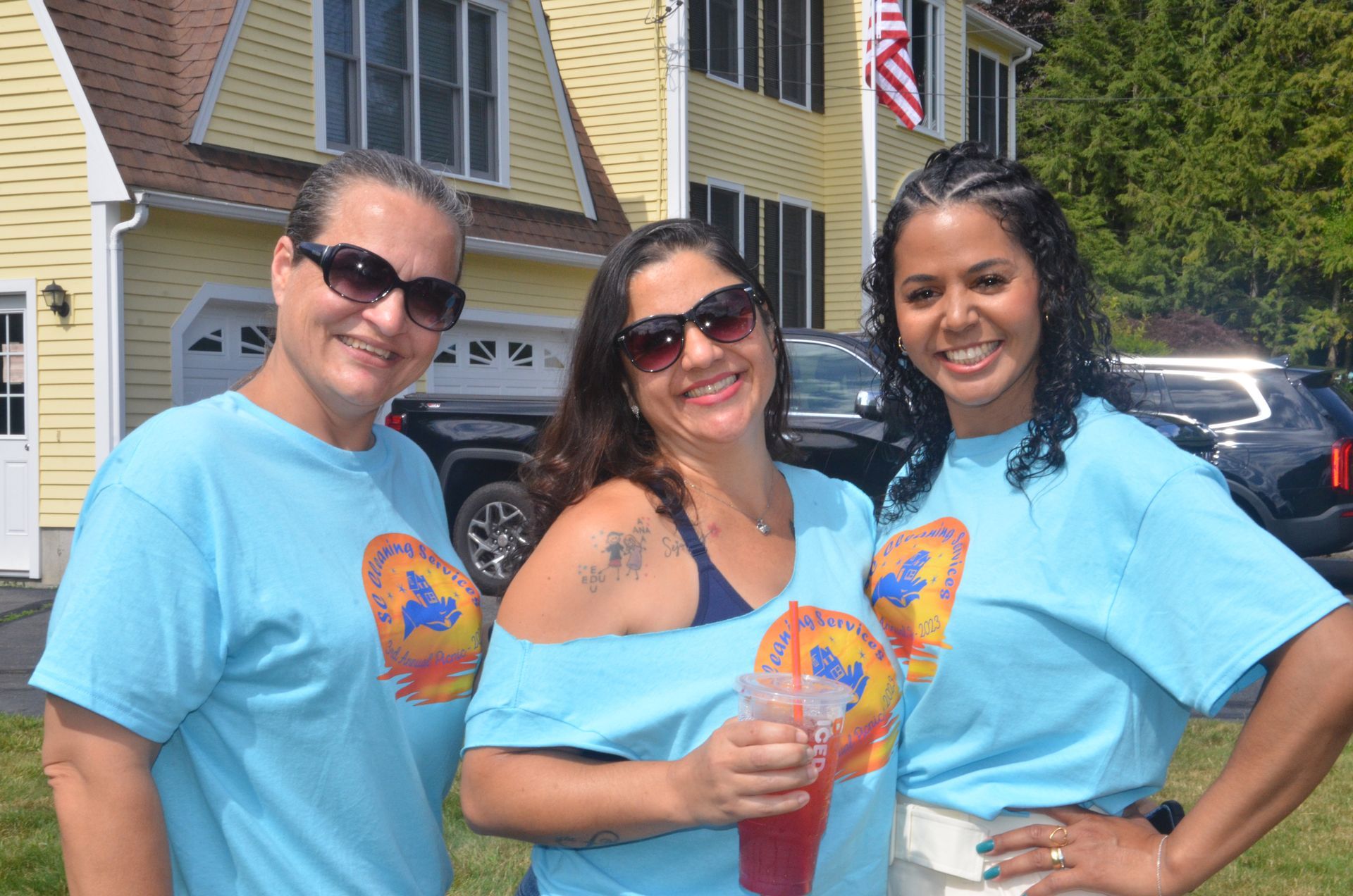 Three women in blue shirts smiling outside a yellow house; they wear sunglasses.