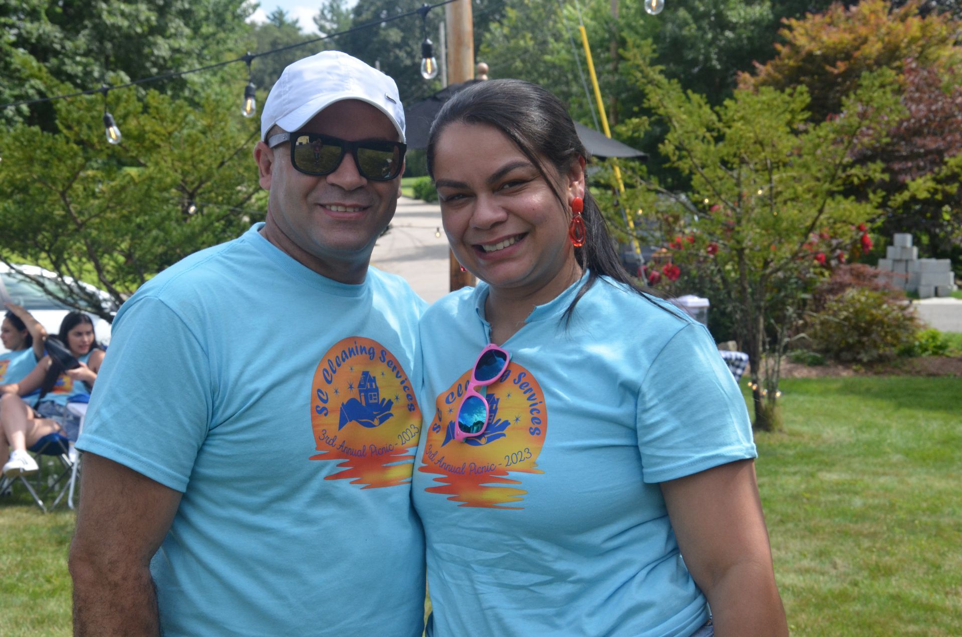Man and woman in blue t-shirts smile, posing outdoors in front of trees, with sunglasses on top of woman's shirt.