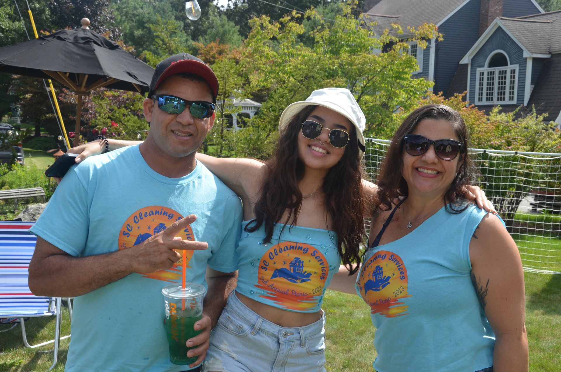 Three people wearing blue t-shirts pose outdoors. Man on left holds drink and makes peace sign.