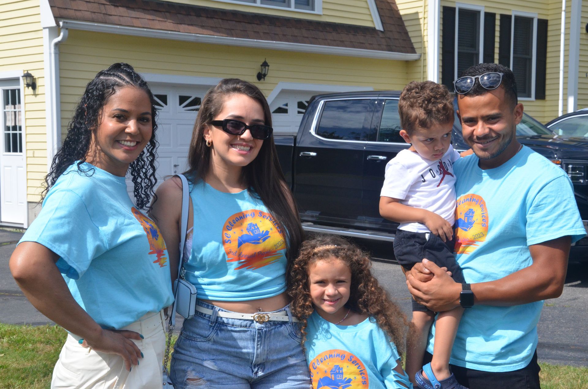 Family of five in matching blue shirts posing in front of a yellow house.