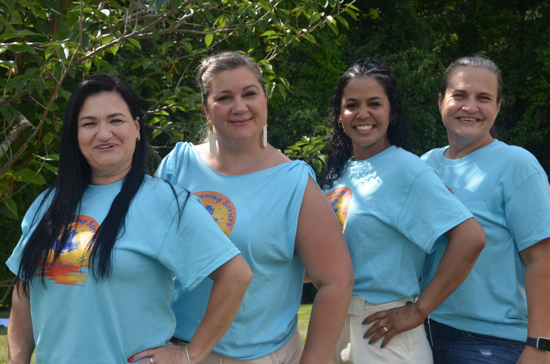 Four women in matching blue shirts smile outdoors, arms on hips.