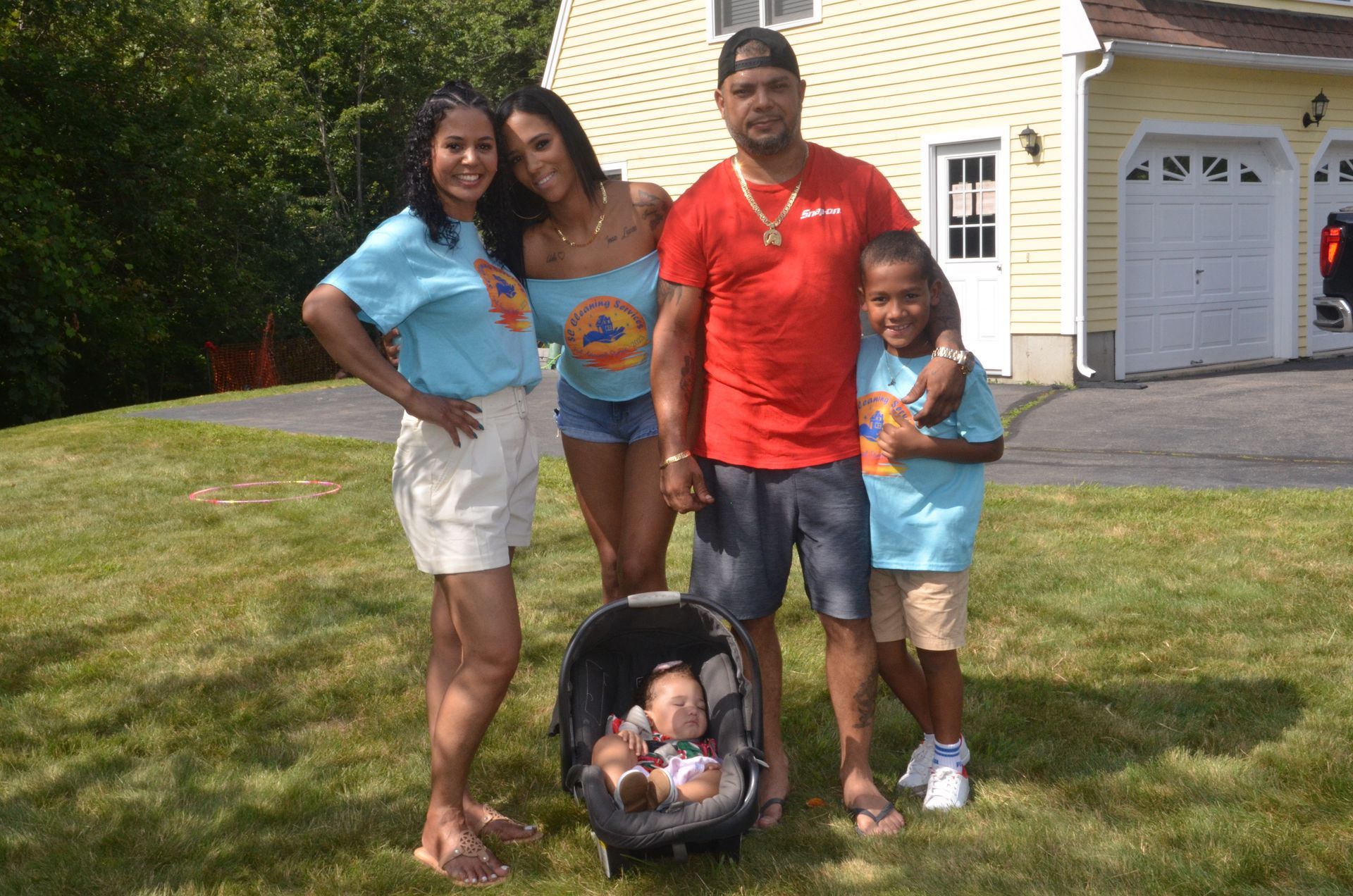 Family of five poses outdoors in front of a house; adults wearing various blue and red clothing, child in car seat.