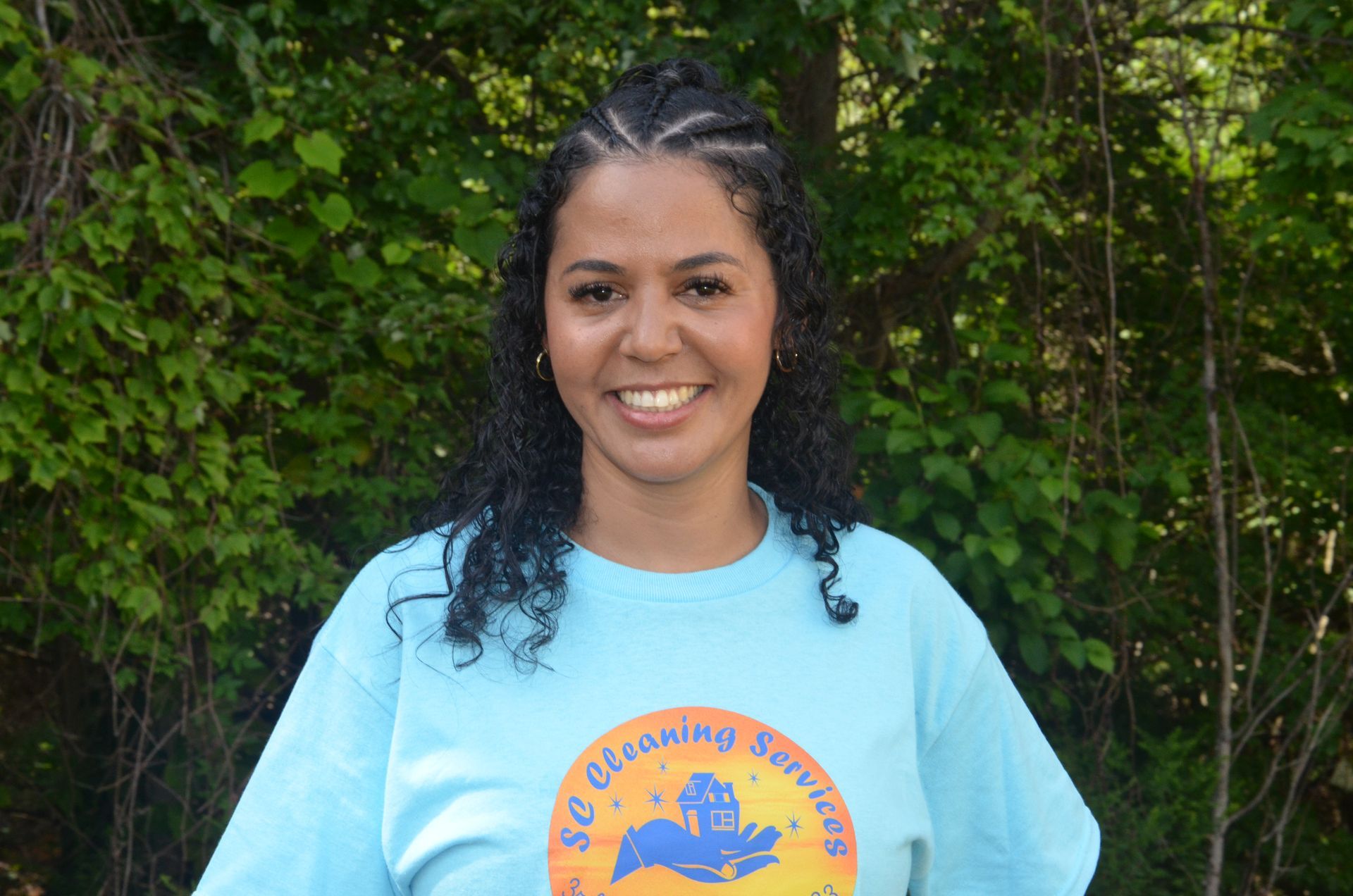 Woman in a light blue shirt with logo, smiling, braids, standing outdoors in front of green foliage.
