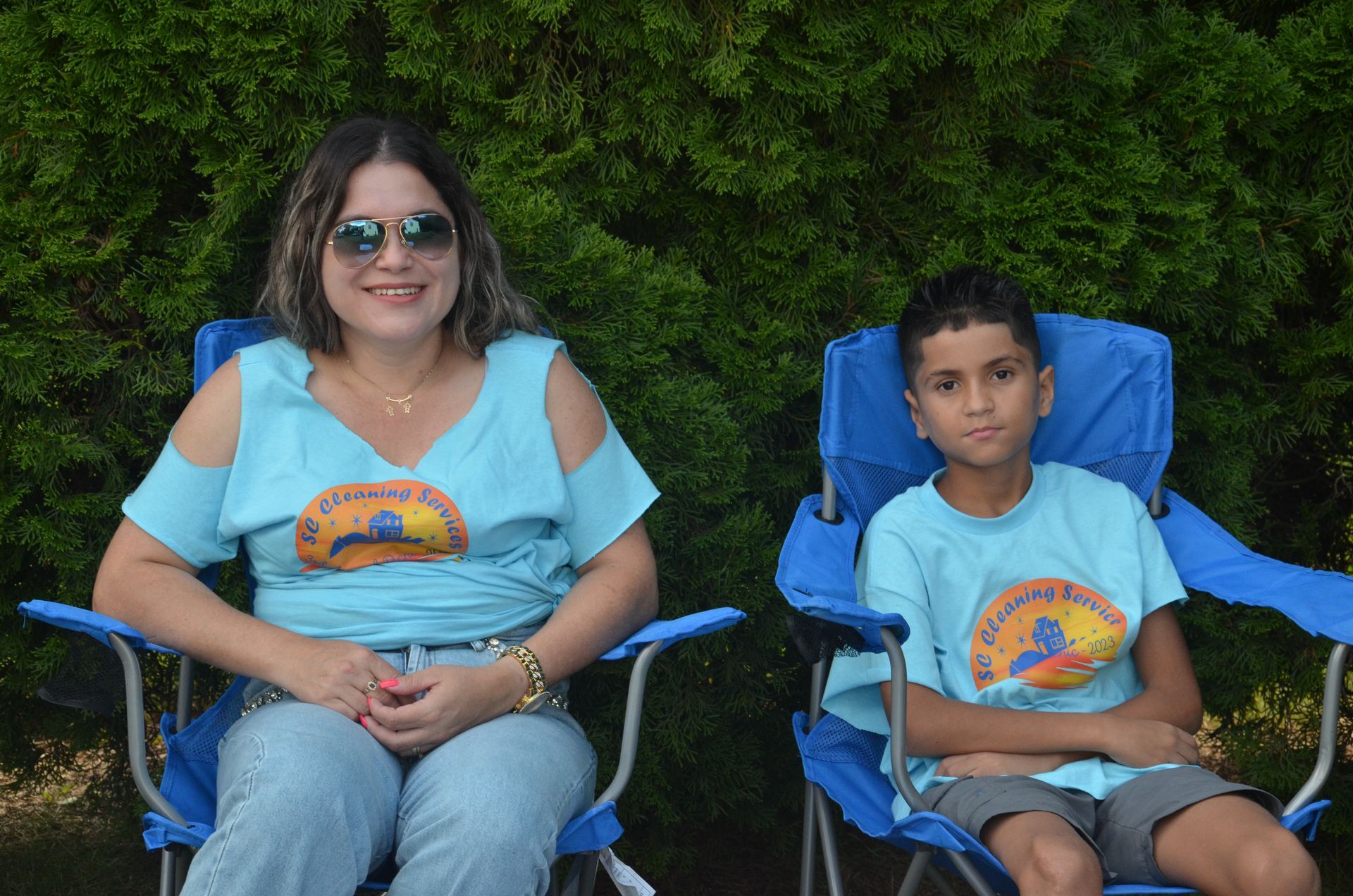 Woman and boy in blue chairs, wearing matching blue t-shirts, smiling, outside.