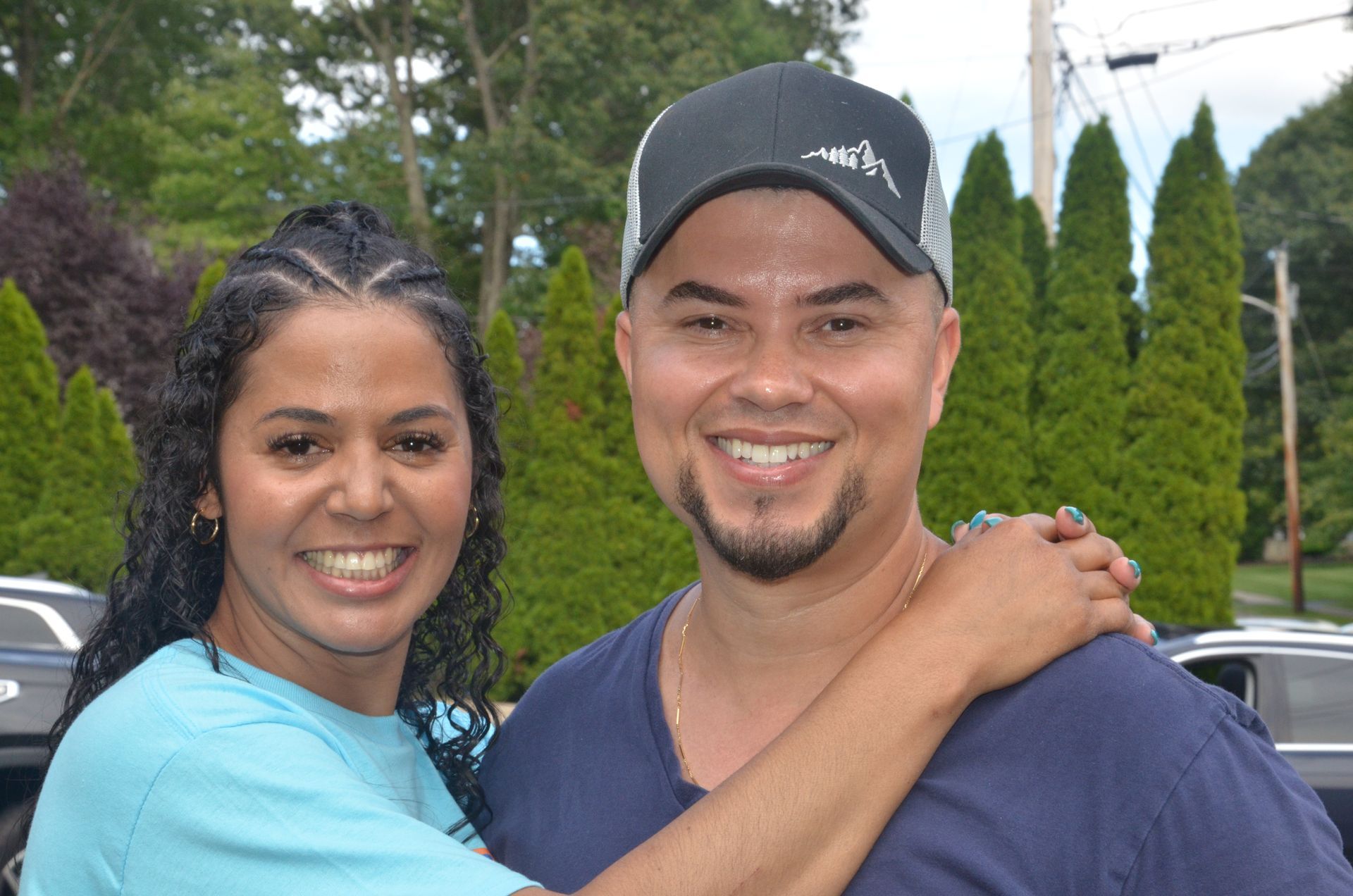 Smiling couple poses outdoors; woman has braided hair, man wears hat and navy shirt.