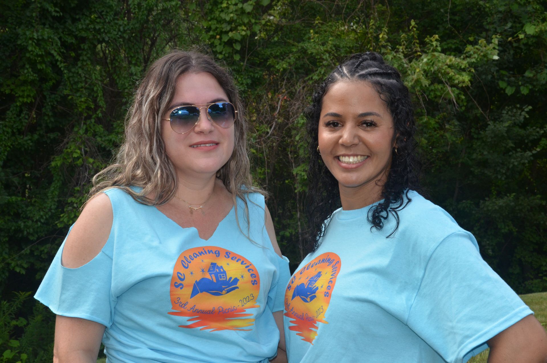 Two women in blue t-shirts, smiling in front of green foliage. One wears sunglasses.