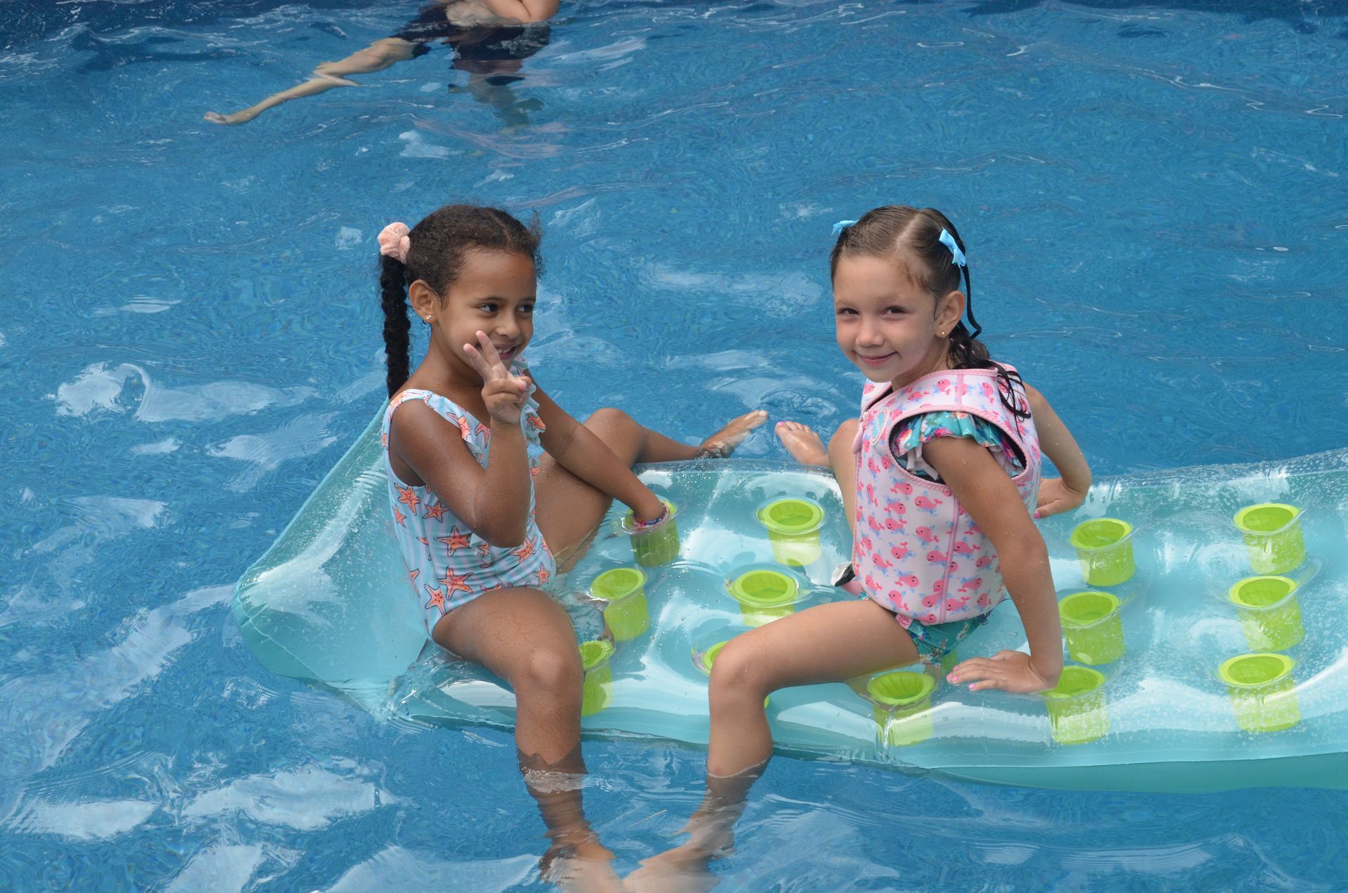 Two girls in swimsuits on a raft in a pool, one making a peace sign.