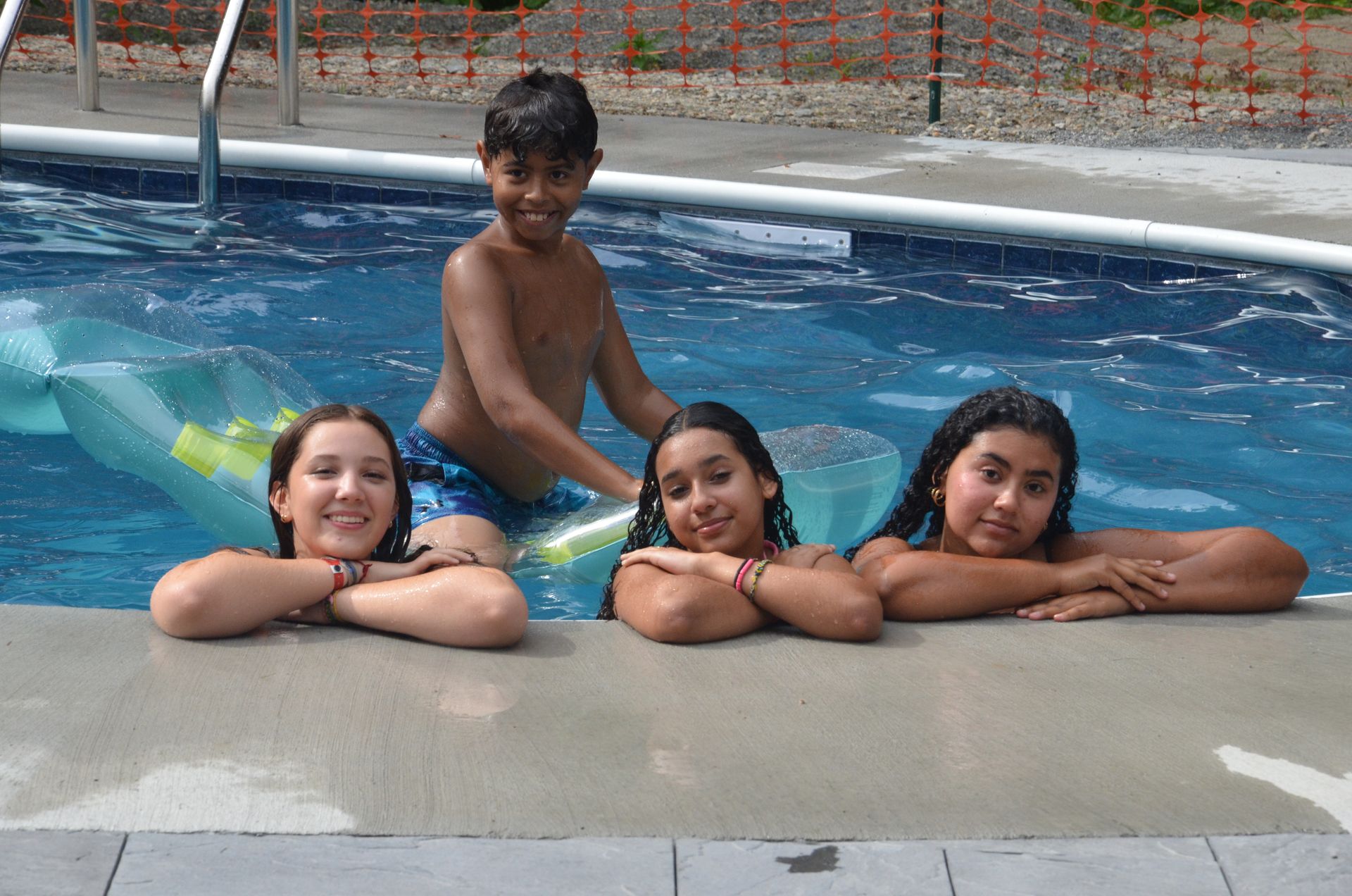 Four children in a pool, smiling. One boy stands in the water, girls resting arms on the edge. Sunny outdoor setting.