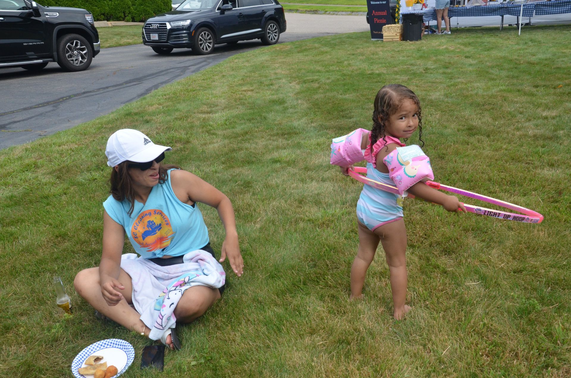 Woman sitting on grass watches a young girl in a swimsuit wearing arm floaties playing with a hula hoop.