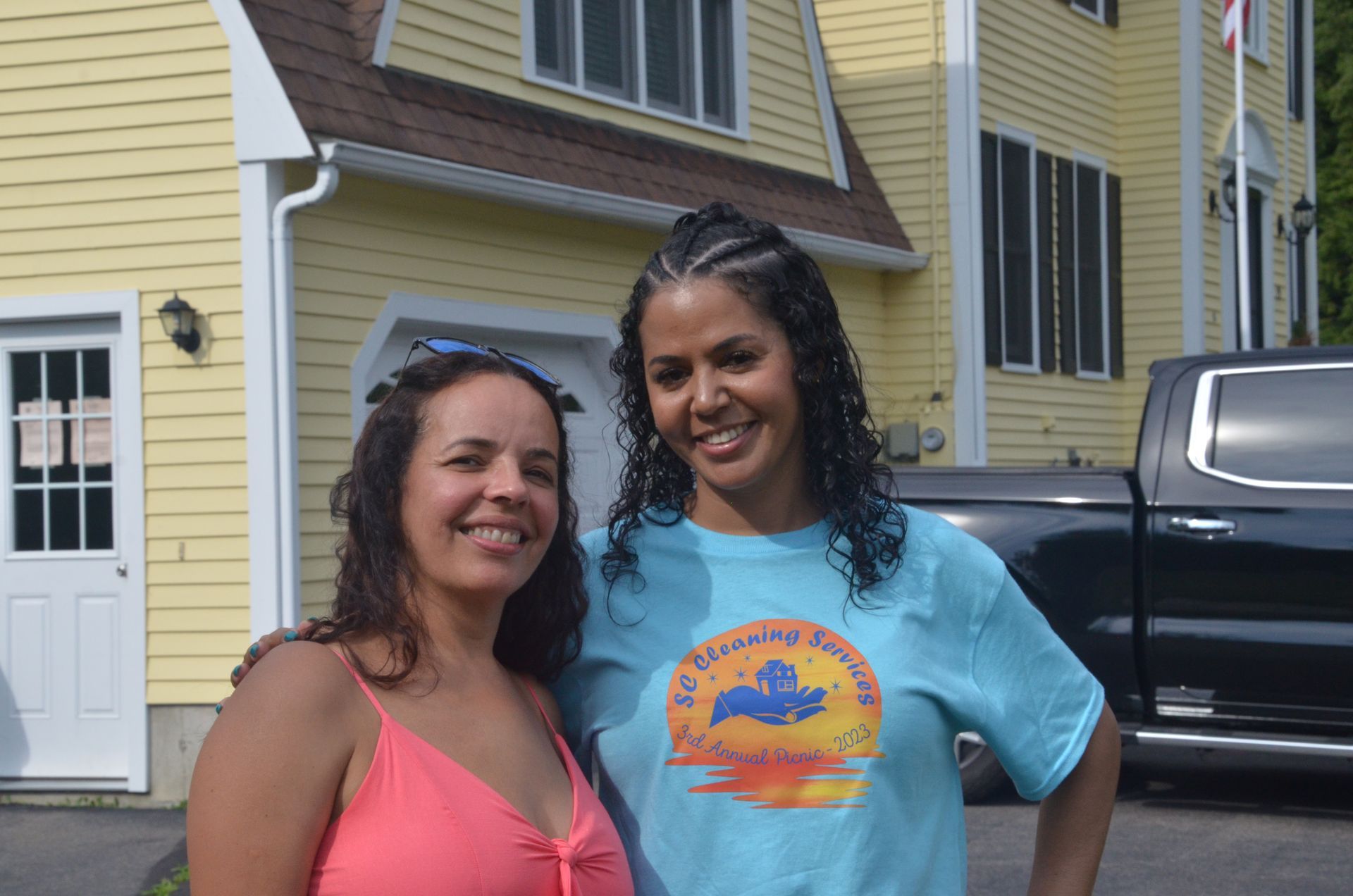 Two women smiling in front of a yellow house. One wears a pink top, the other a blue t-shirt.