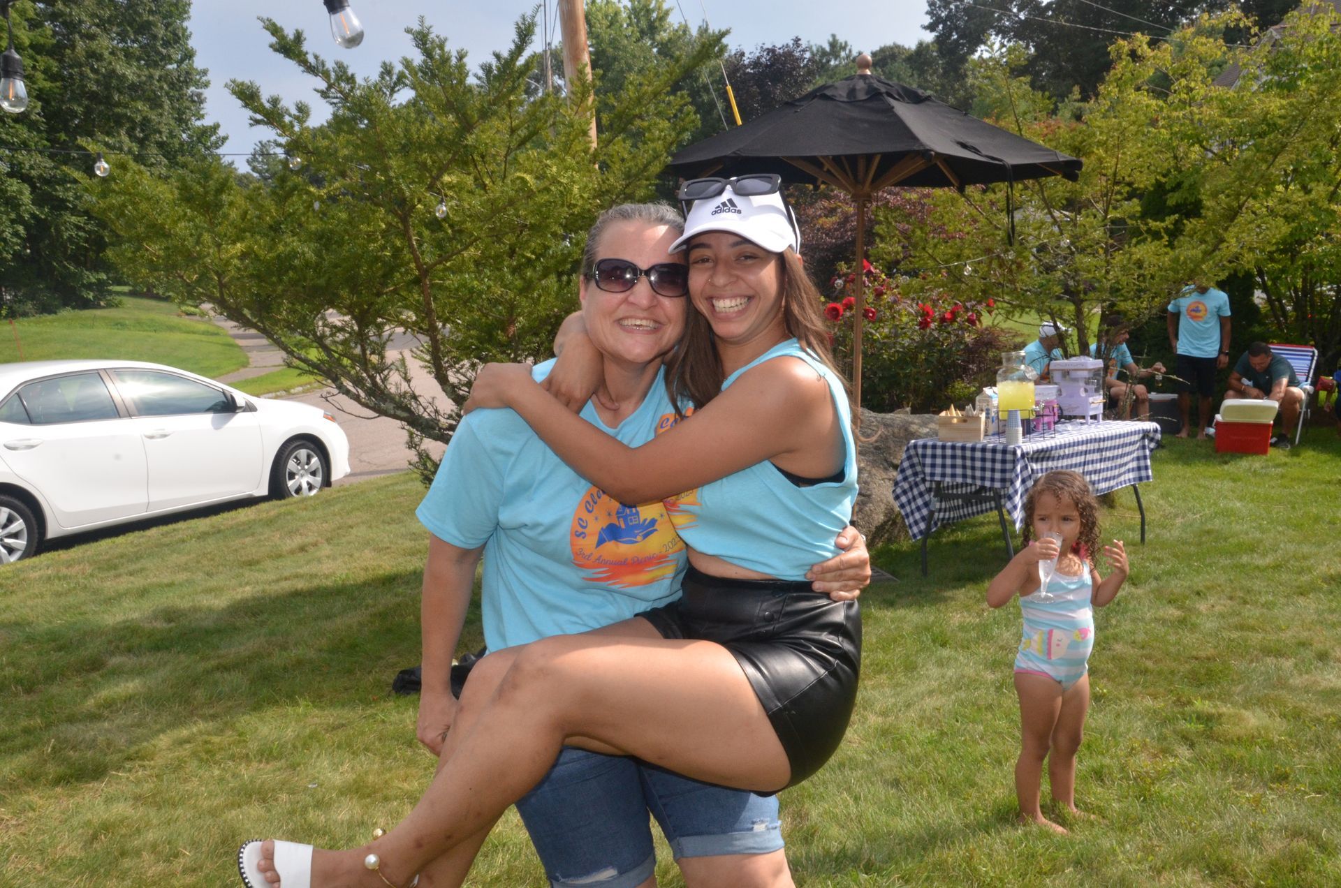 Woman carrying a young woman in front of party setting. Both smiling, wearing light blue shirts.