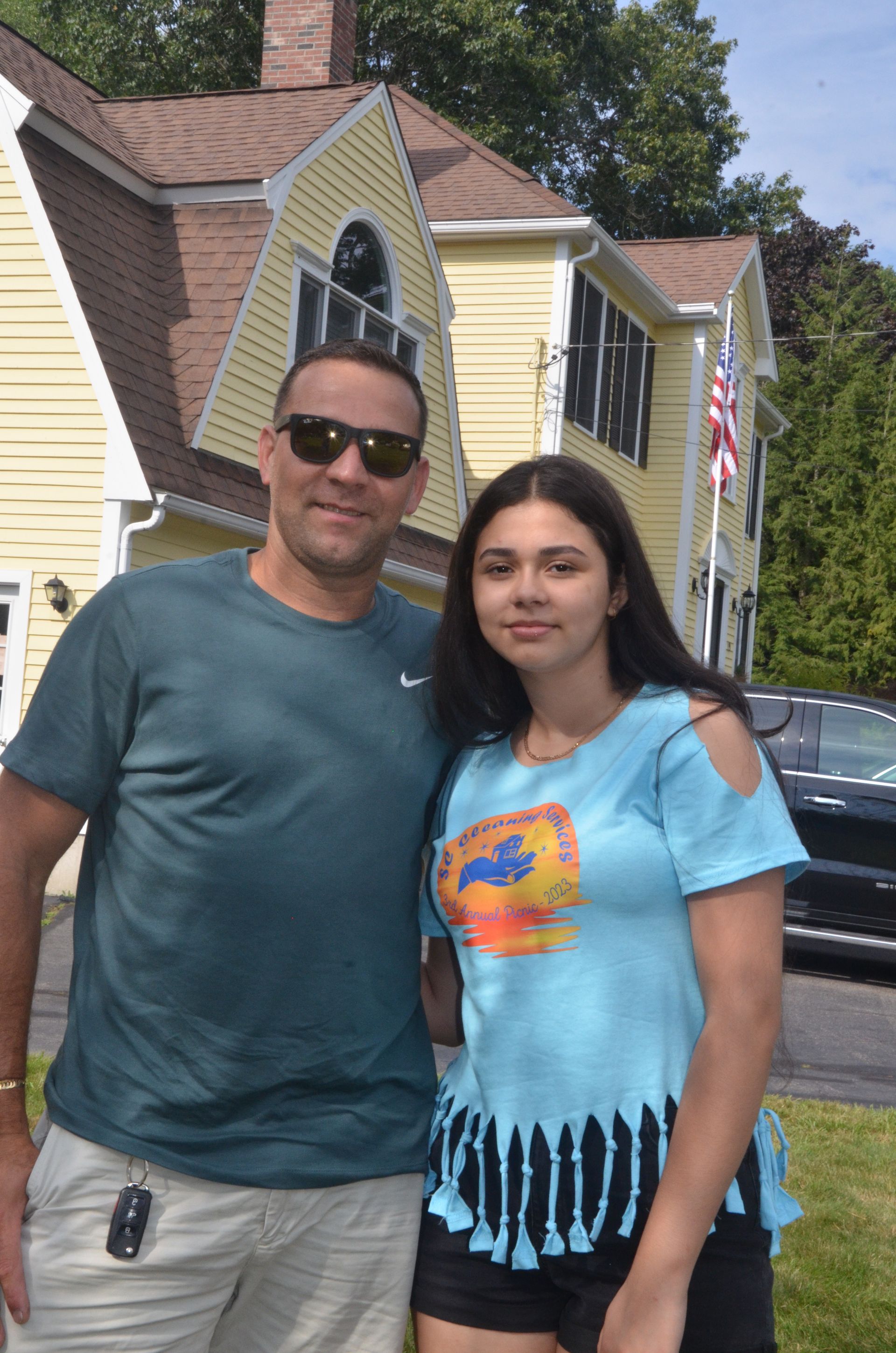 Man with sunglasses and a young woman stand in front of a yellow house; they smile, sunny day.
