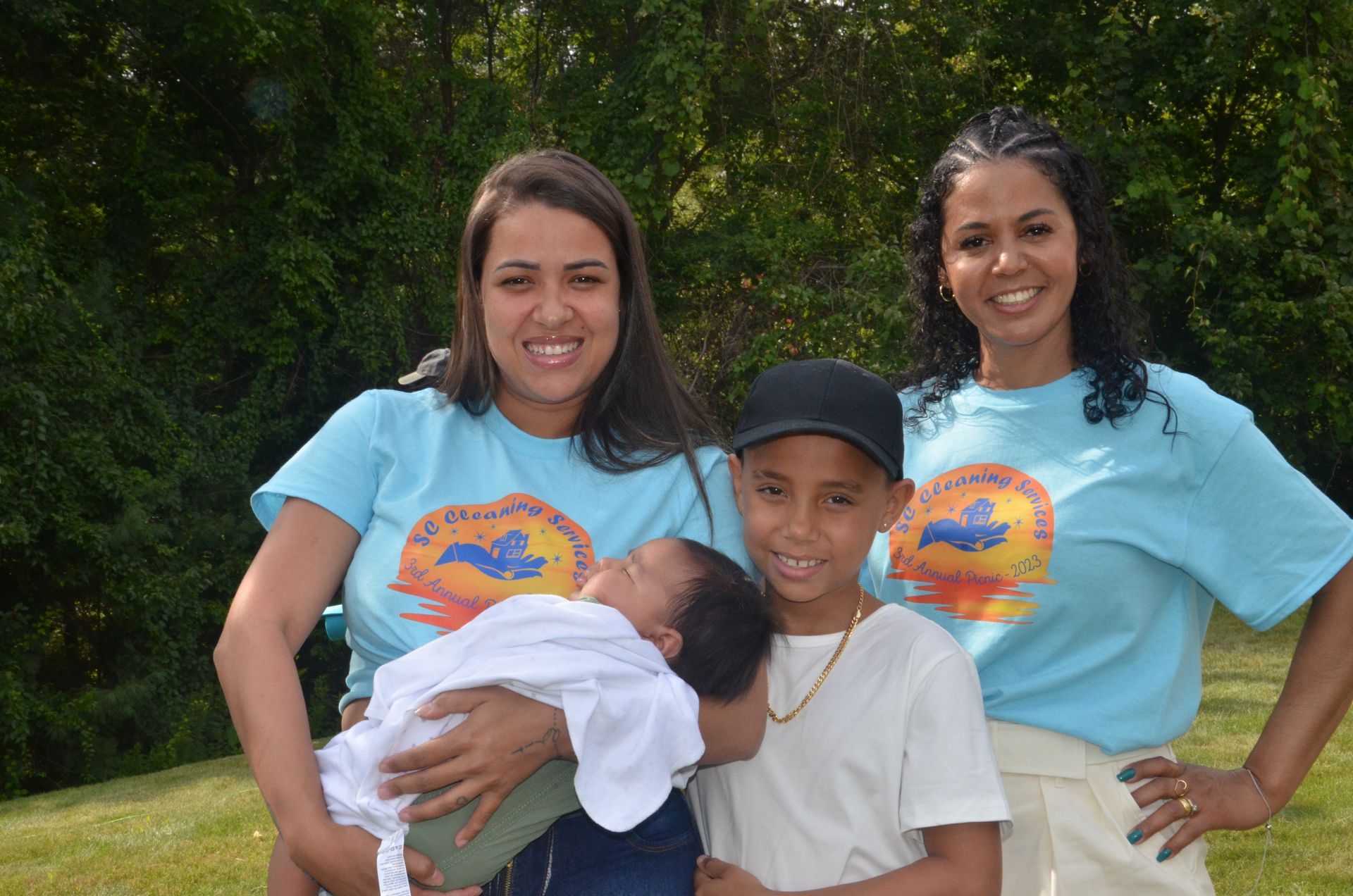 Two women and two children outdoors; women in blue shirts, one holding a baby, other child wearing a cap.