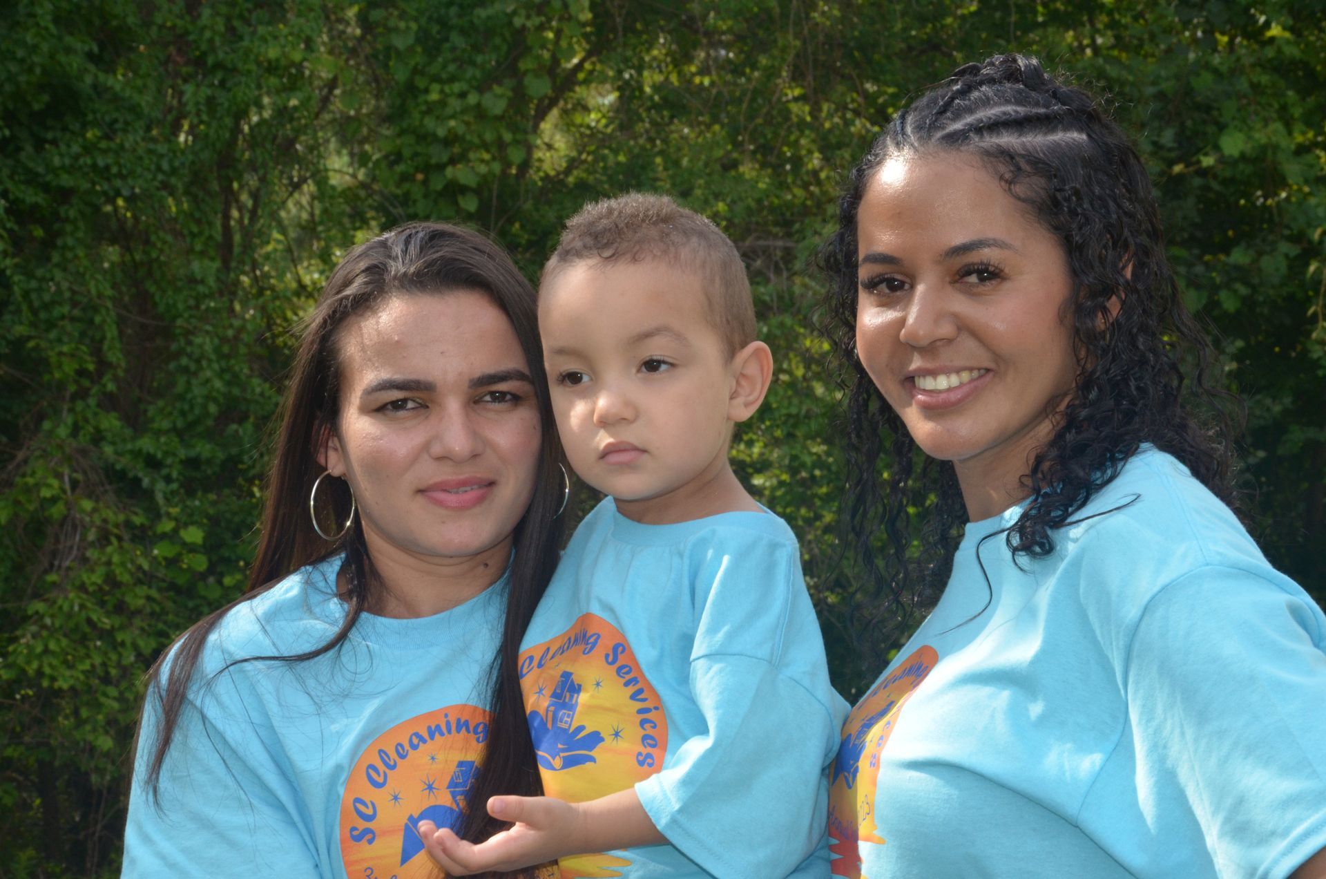 Three people wearing light blue shirts stand outside; two women flank a young child.