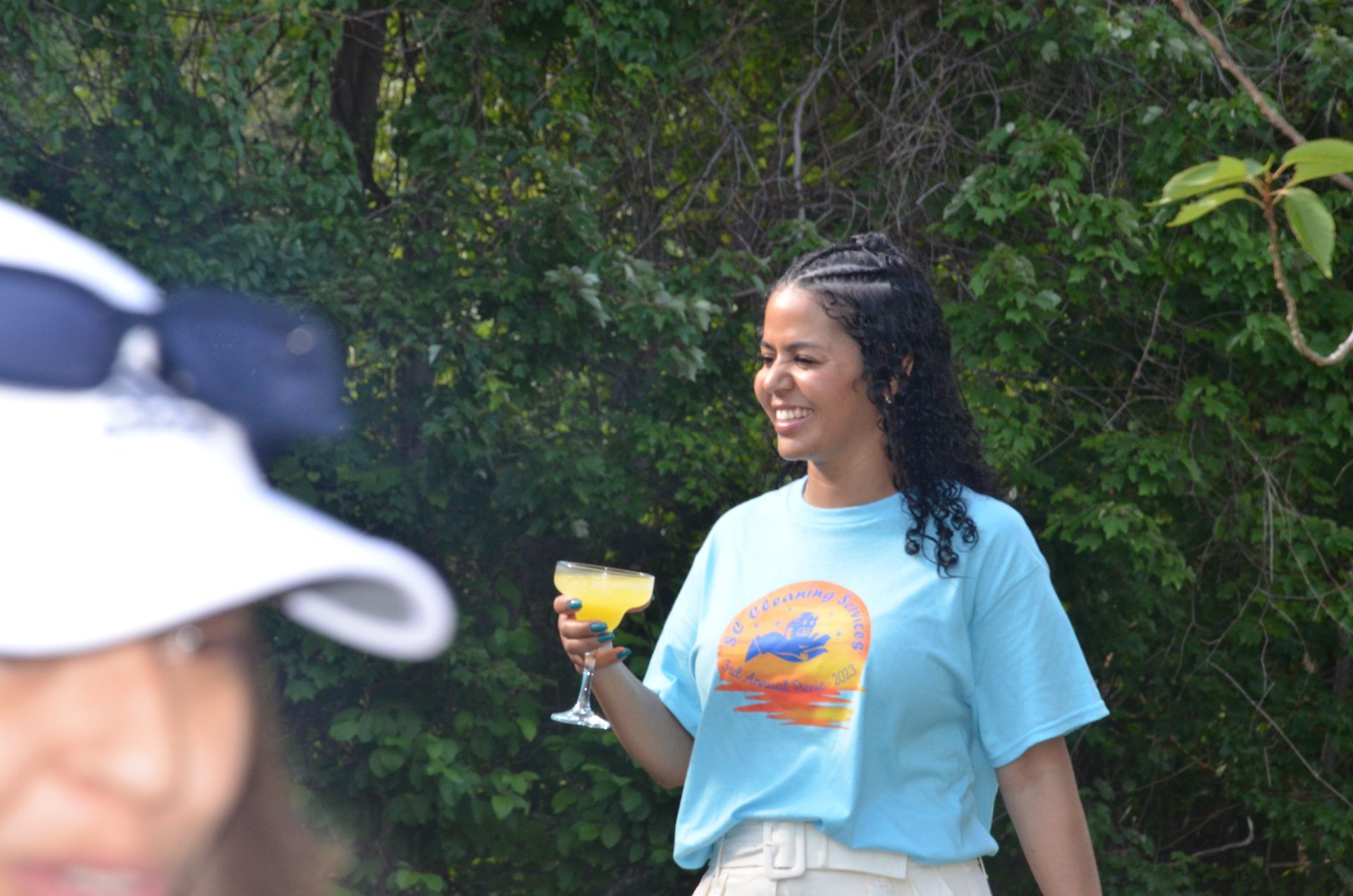 Woman with a cocktail smiles outdoors, wearing a blue shirt and white pants. A white hat is in the foreground.