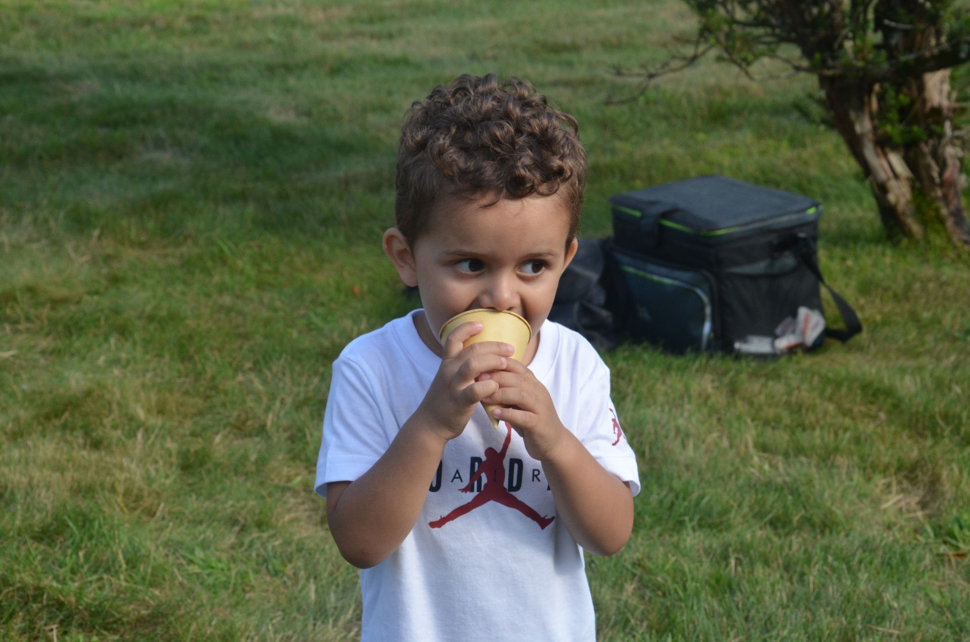 Young child eating a snack outdoors, with curly hair, white t-shirt, and green grass background.